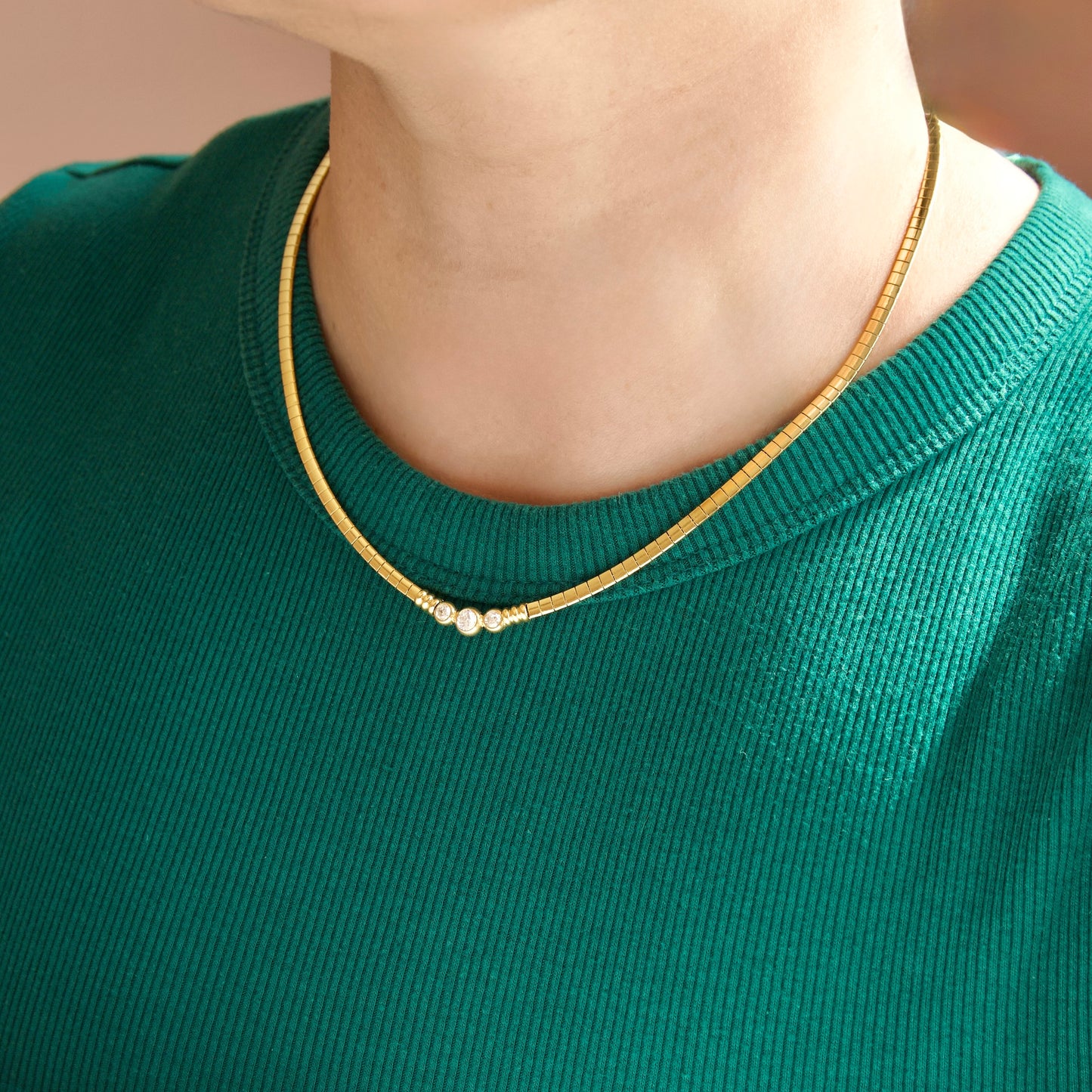 A close-up of a person wearing a delicate gold chain necklace with three small white CZ stones at the center, styled over a green ribbed knit top.