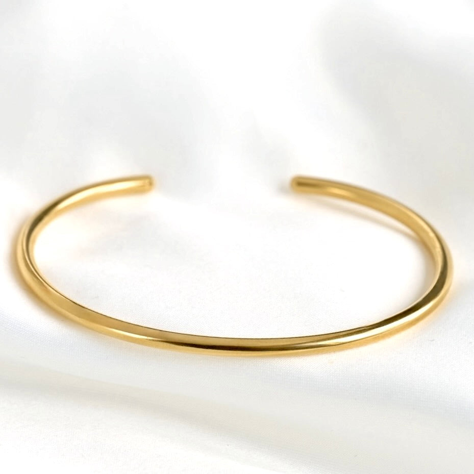 A thin polished gold open cuff bracelet resting on a soft, white textured fabric background.