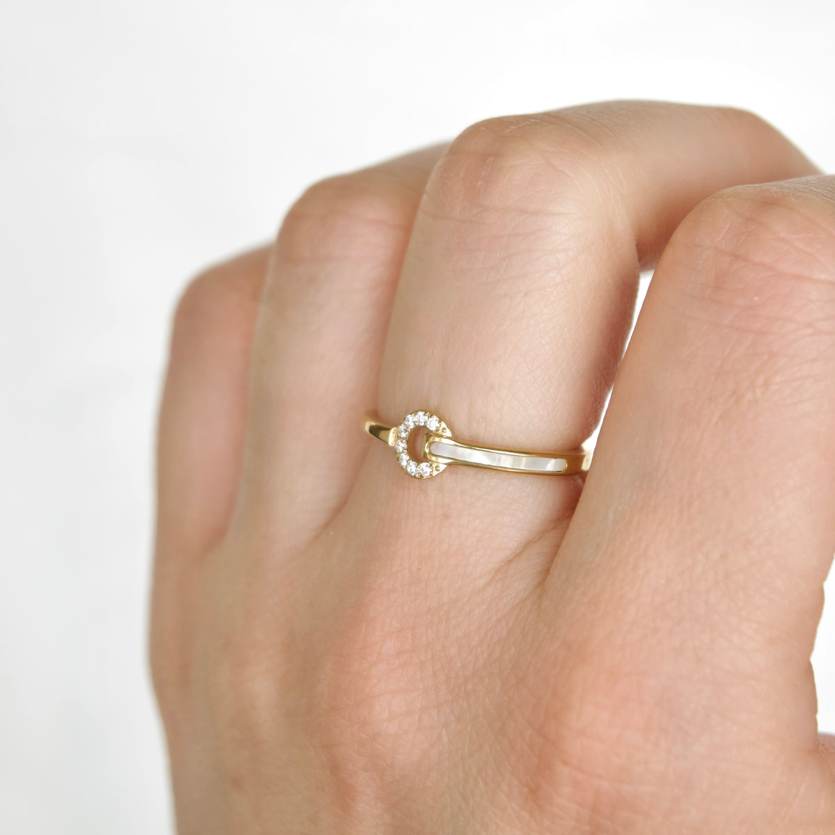 A close-up of a hand wearing a Delicate gold ring with a small circular diamond halo and shell inlay on a plain light background.