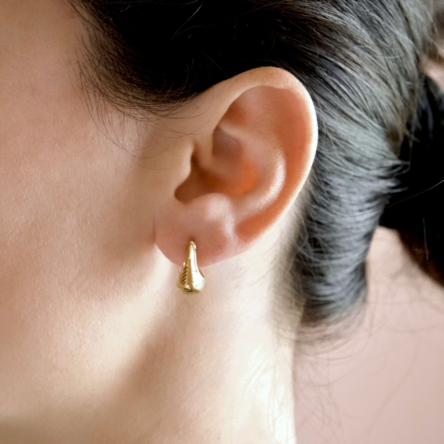 A close-up of a person's ear wearing a small gold huggie hoop earring with a textured teardrop drop.