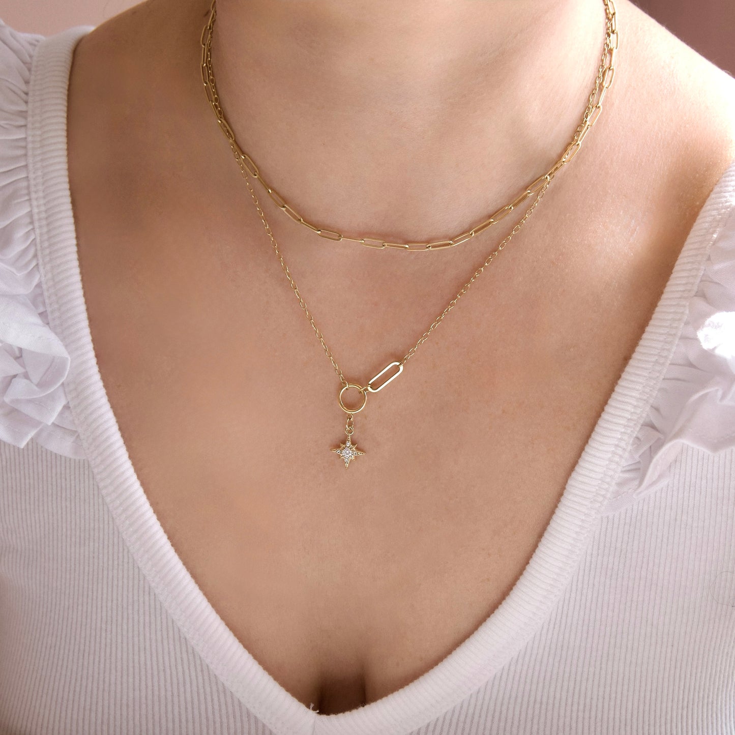 A close-up of a model's neck and chest wearing a double-layered gold necklace. The top layer is a thicker paperclip chain, and the bottom layer is a delicate chain with a small gold paperclip ring and a tiny clear crystal star charm.