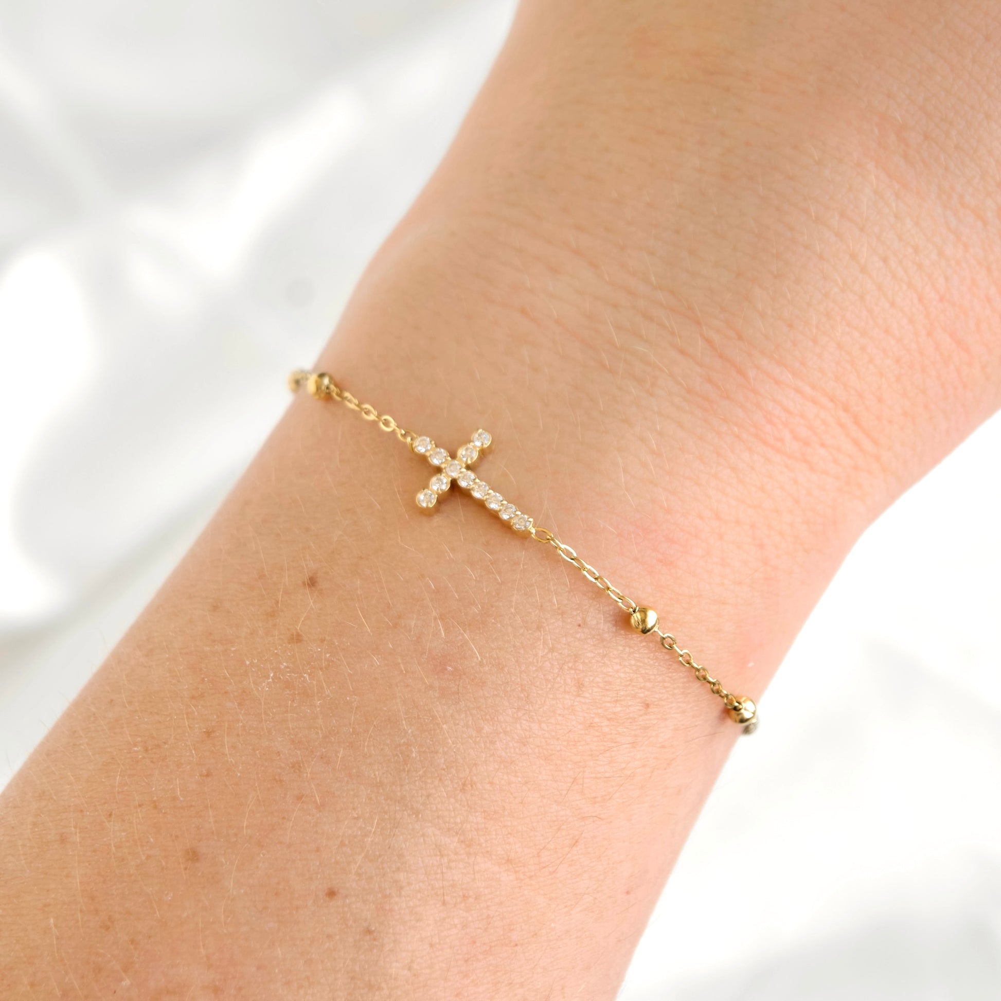 A delicate gold chain bracelet with a small cross charm set with clear stones, shown worn on a person's wrist against a soft white background.