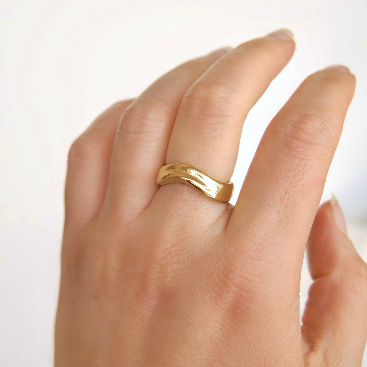 A close-up of a hand wearing a polished yellow gold band ring with a wavy, organic textured design, set against a clean white background.