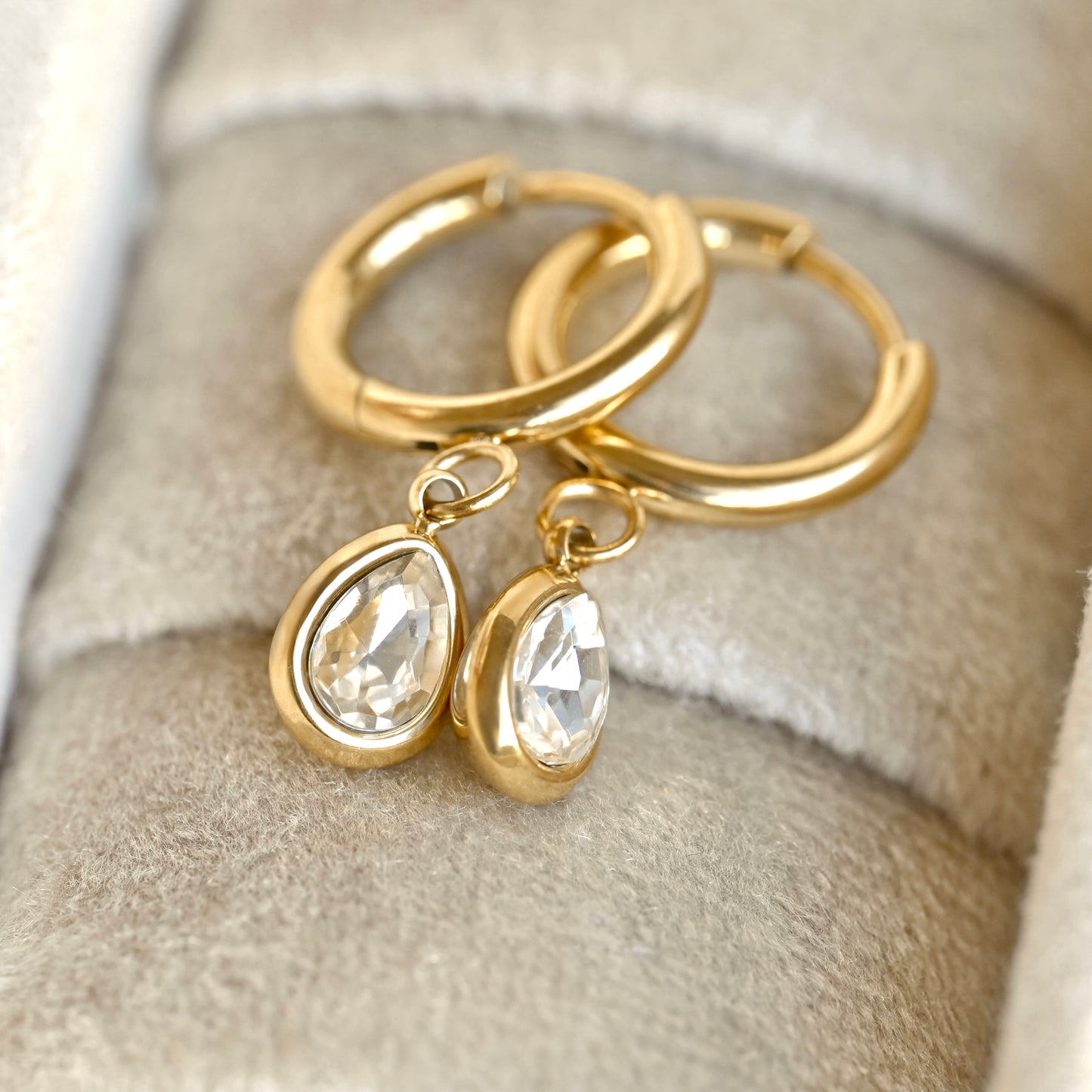 A pair of gold hoop earrings with waterdrop clear crystal drops, displayed on a soft beige velvet jewelry box.