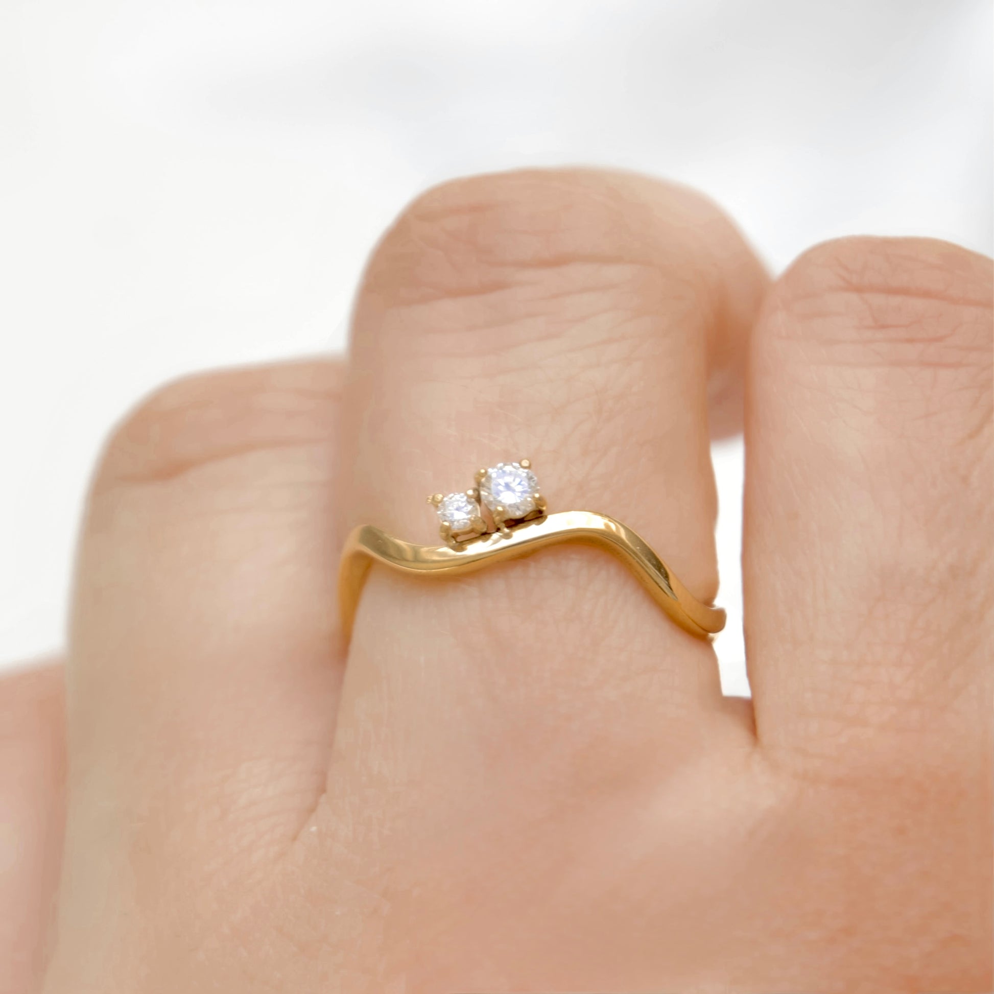 A close-up of a gold wave ring with two small clear gemstones, worn on a finger.