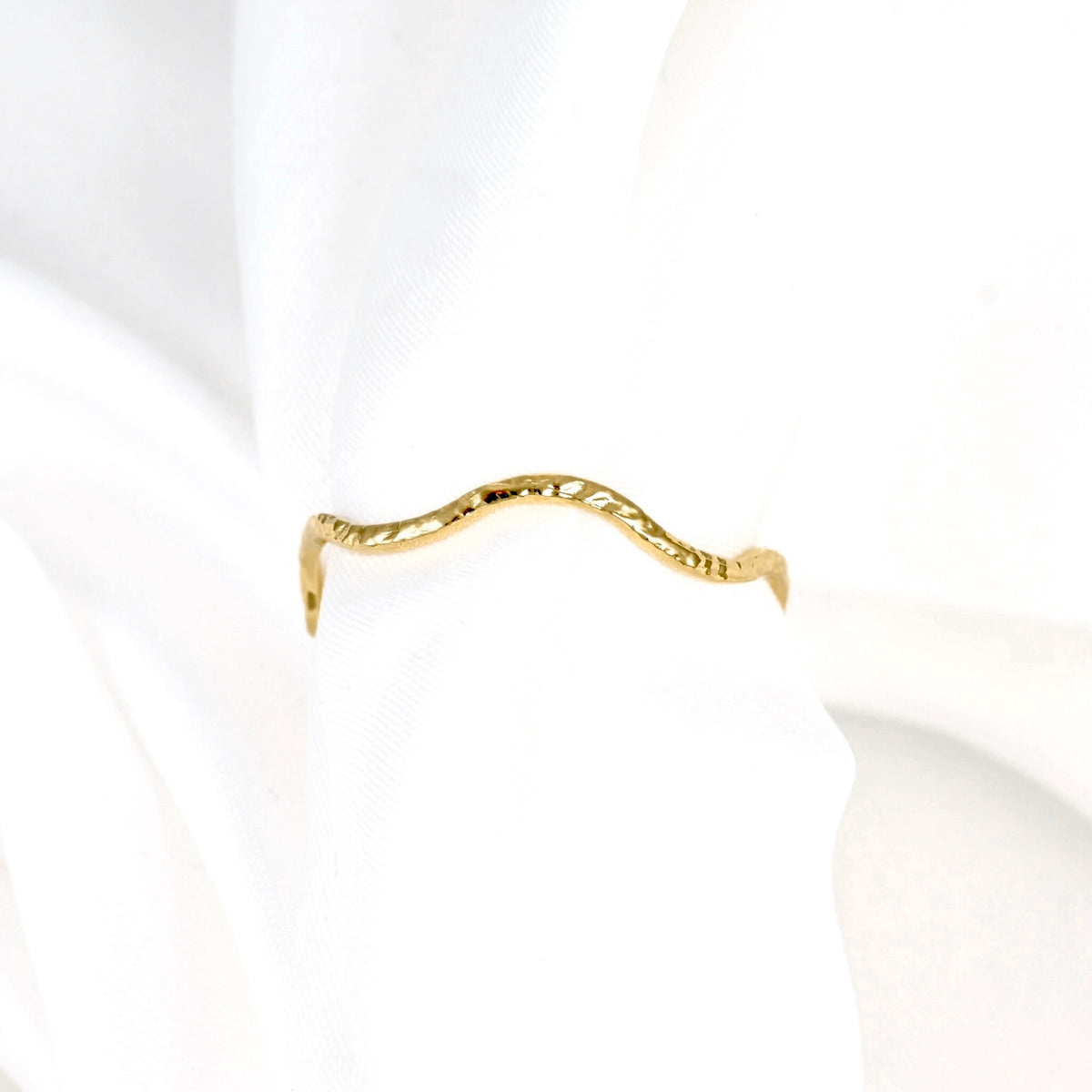 A delicate, wavy gold ring with a textured, hammered finish, displayed on a soft white fabric background.