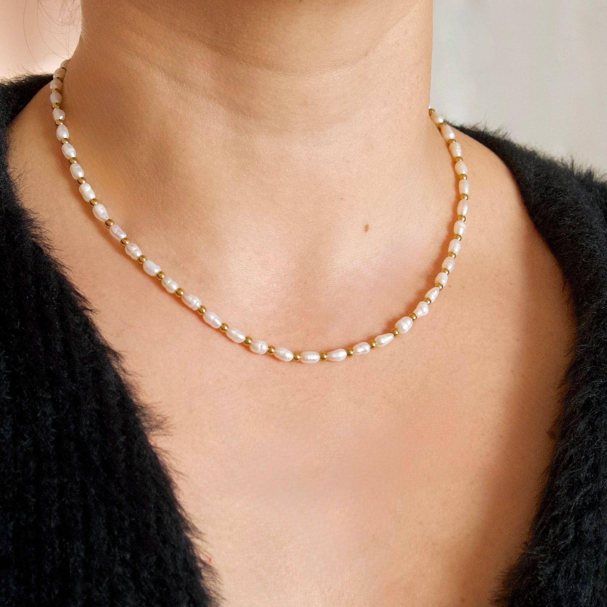 A close-up of a person wearing a delicate gold chain necklace with irregular white freshwater pearls, styled with a black knit top.