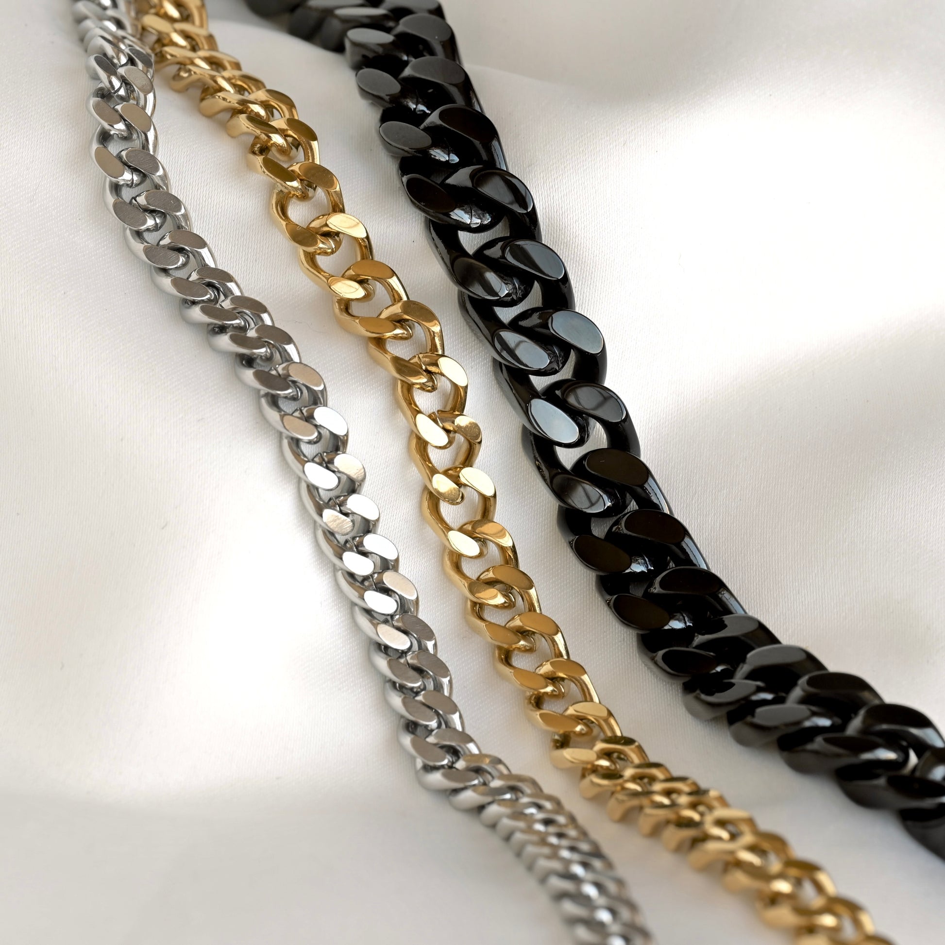 Three chunky curb chains in silver, gold, and black, arranged diagonally on a white satin fabric background.