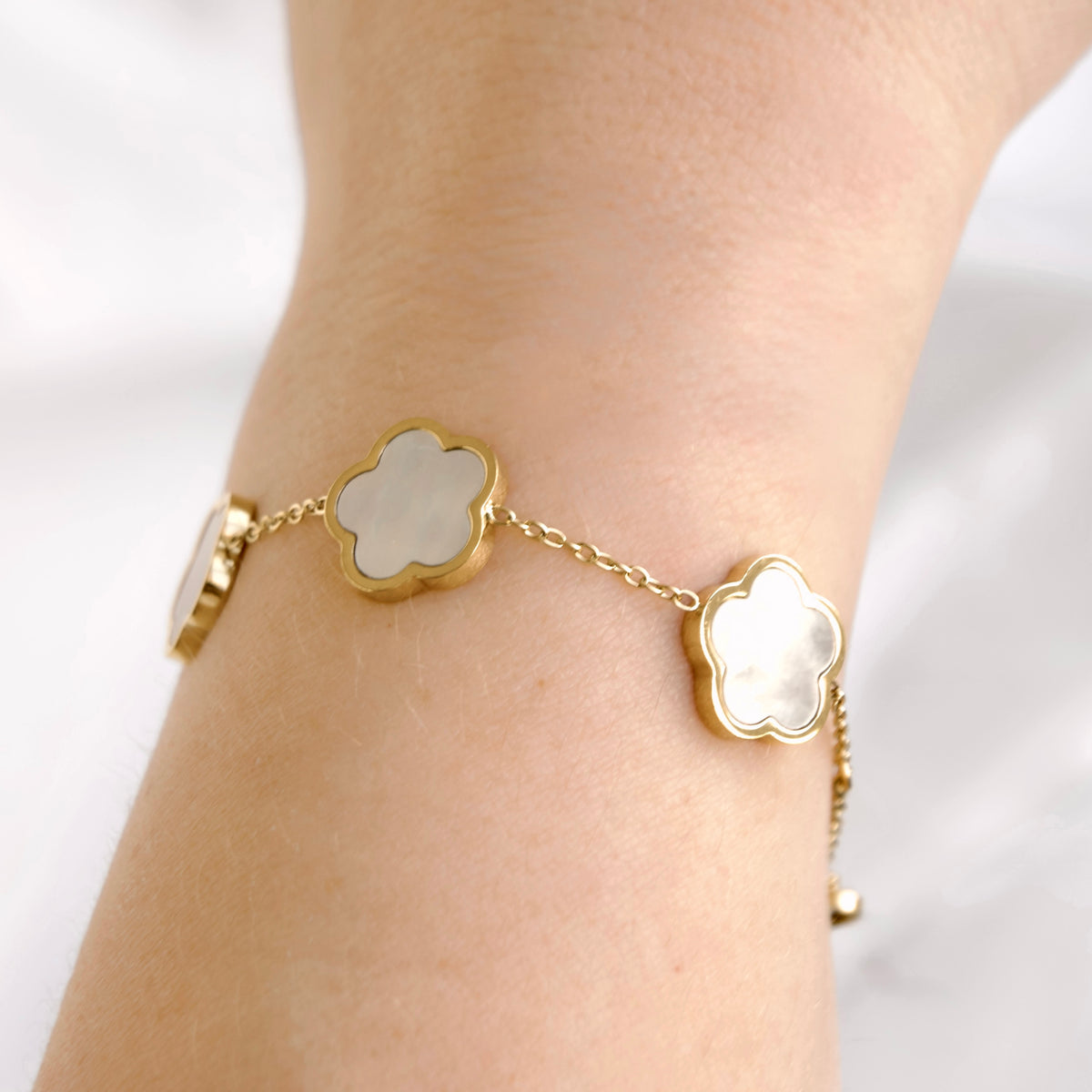 A gold chain bracelet with two clover-shaped mother-of-pearl inlays, shown worn on a person's wrist against a soft white background.