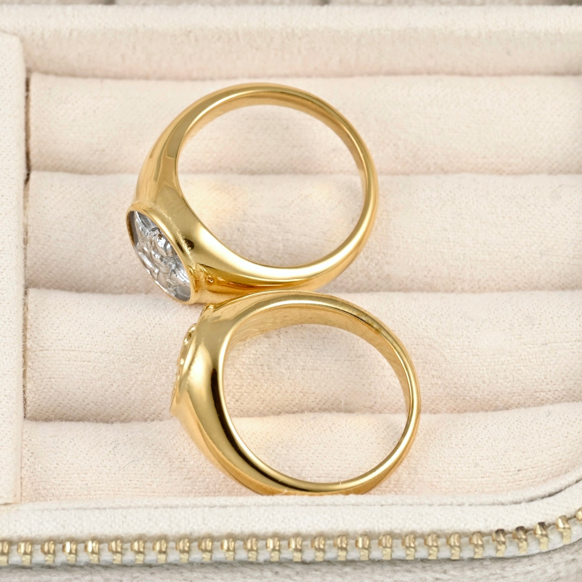 Two chunky gold signet rings , displayed on a beige velvet jewelry box.