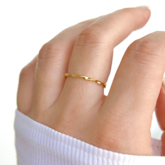 A close-up of a hand wearing a thin, twisted gold band ring, with a white ribbed sleeve visible at the wrist against a bright white background.