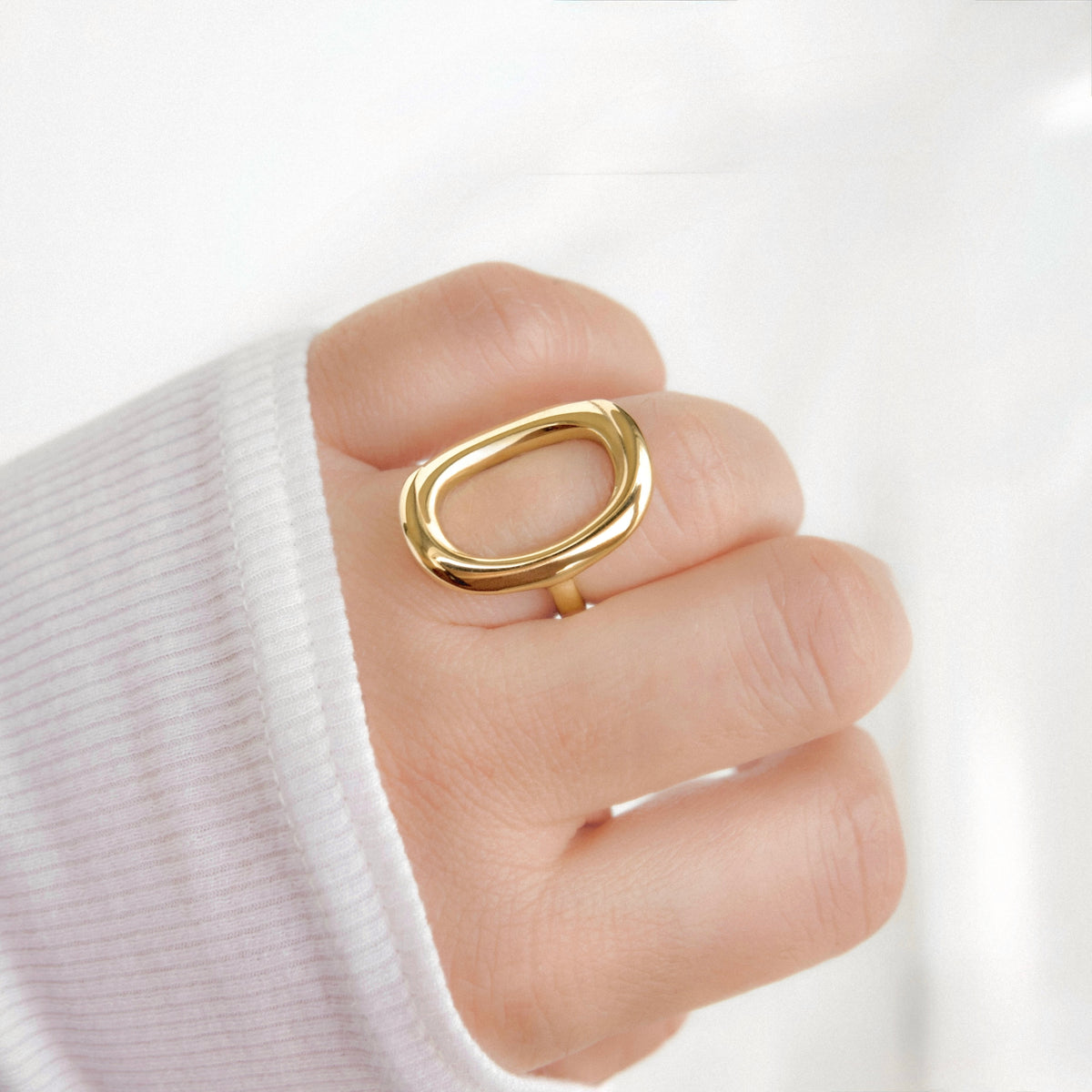 A close-up of a hand wearing a thick, polished gold ring with a rounded, open oval design, styled with a white ribbed knit sleeve.