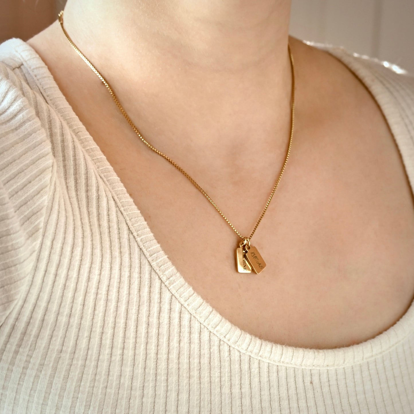 A close-up of a person wearing a delicate gold chain necklace with two small overlapping tag pendants, styled with a cream ribbed scoop-neck top.