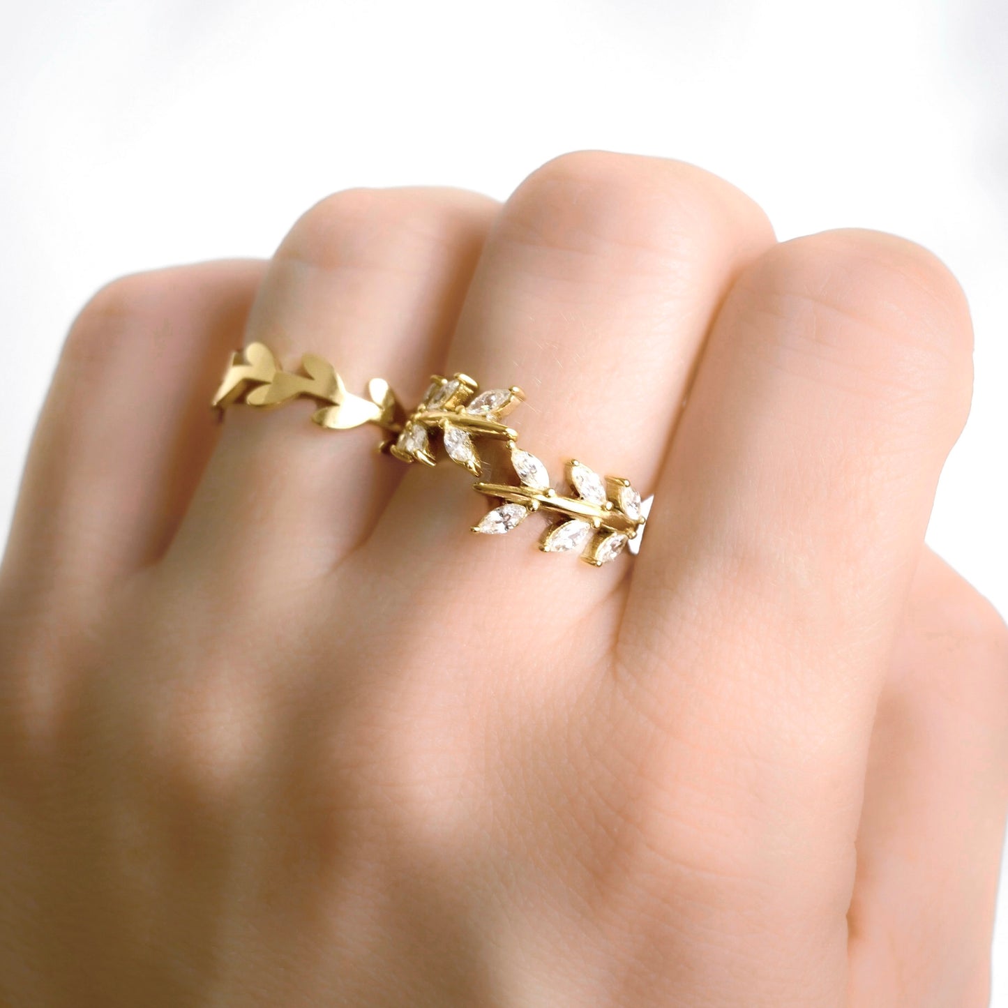 A gold leaf bypass ring with small clear marquise cut stones in middle finger and leaf vine ring on ring finger against a bright white background.