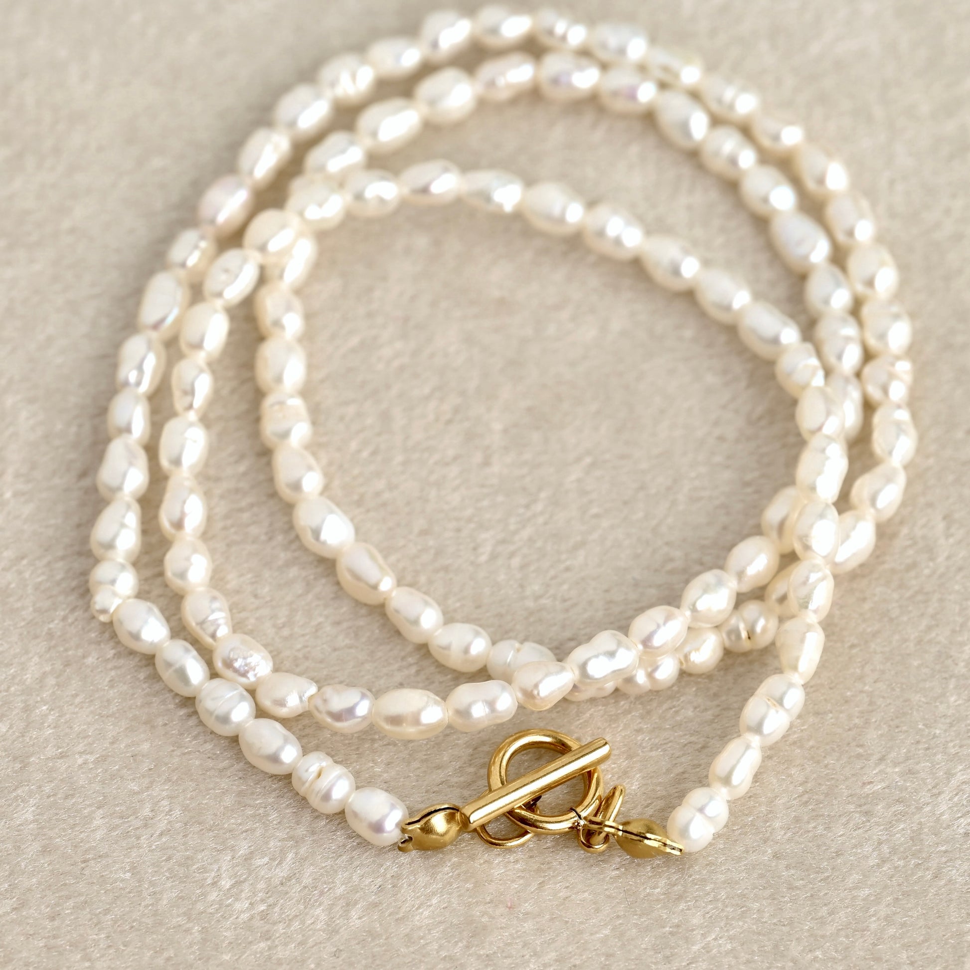 A small baroque pearl necklace with a gold toggle clasp, coiled on a textured beige fabric background.