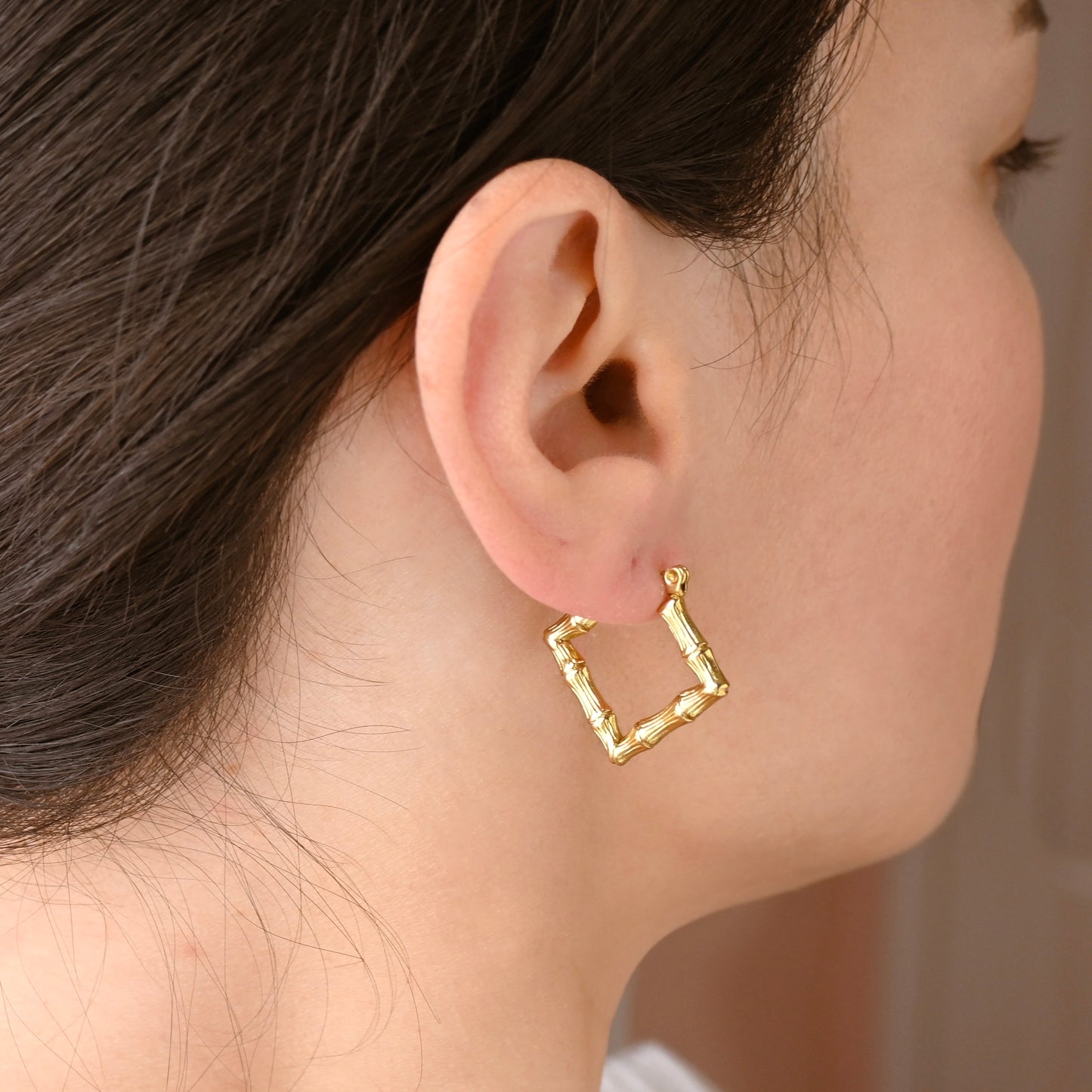 A close-up side profile of a woman wearing a textured  square gold hoop earring.