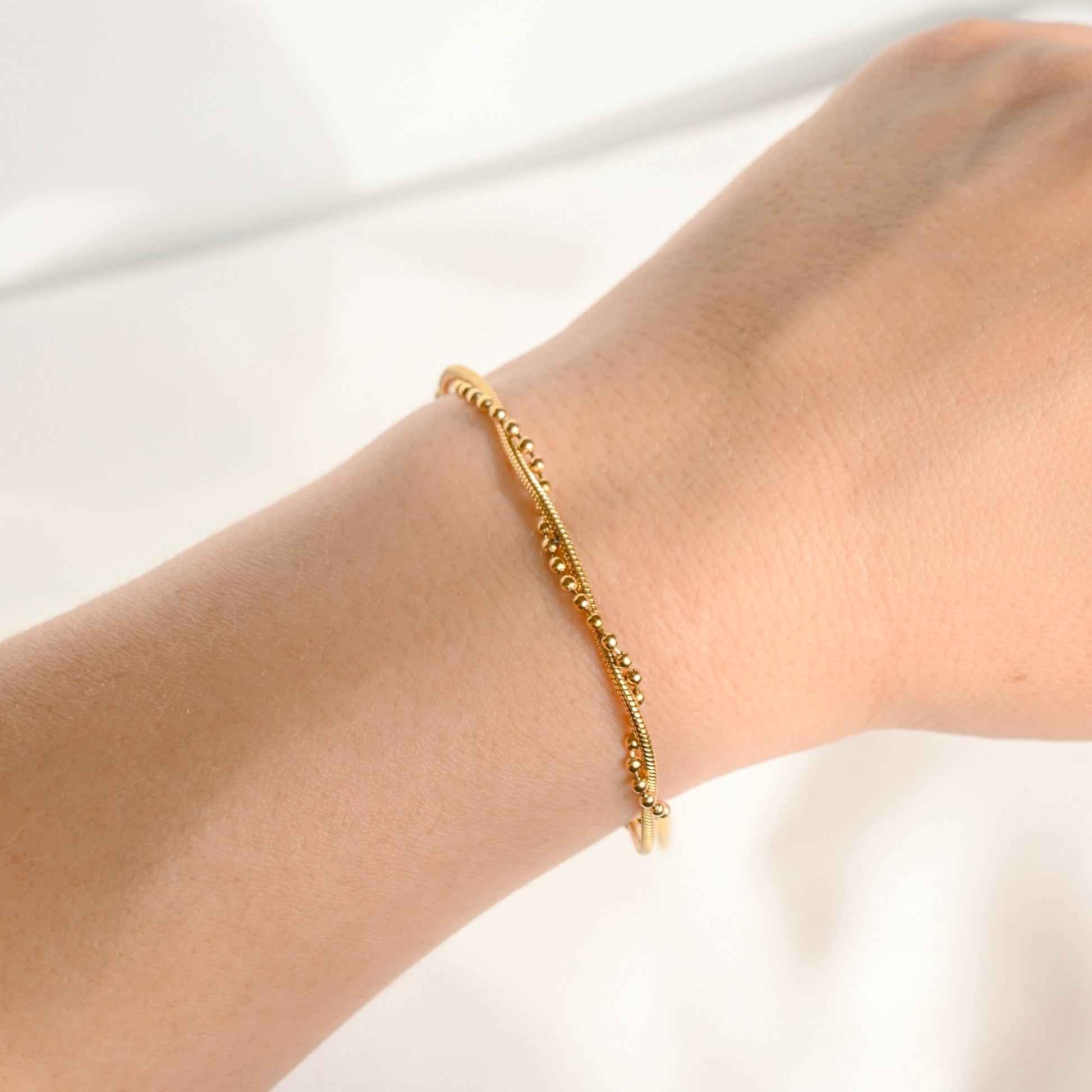 A delicate gold beaded twisted chain bracelet worn on a person's wrist, shown in a close-up shot against a bright, soft white background.