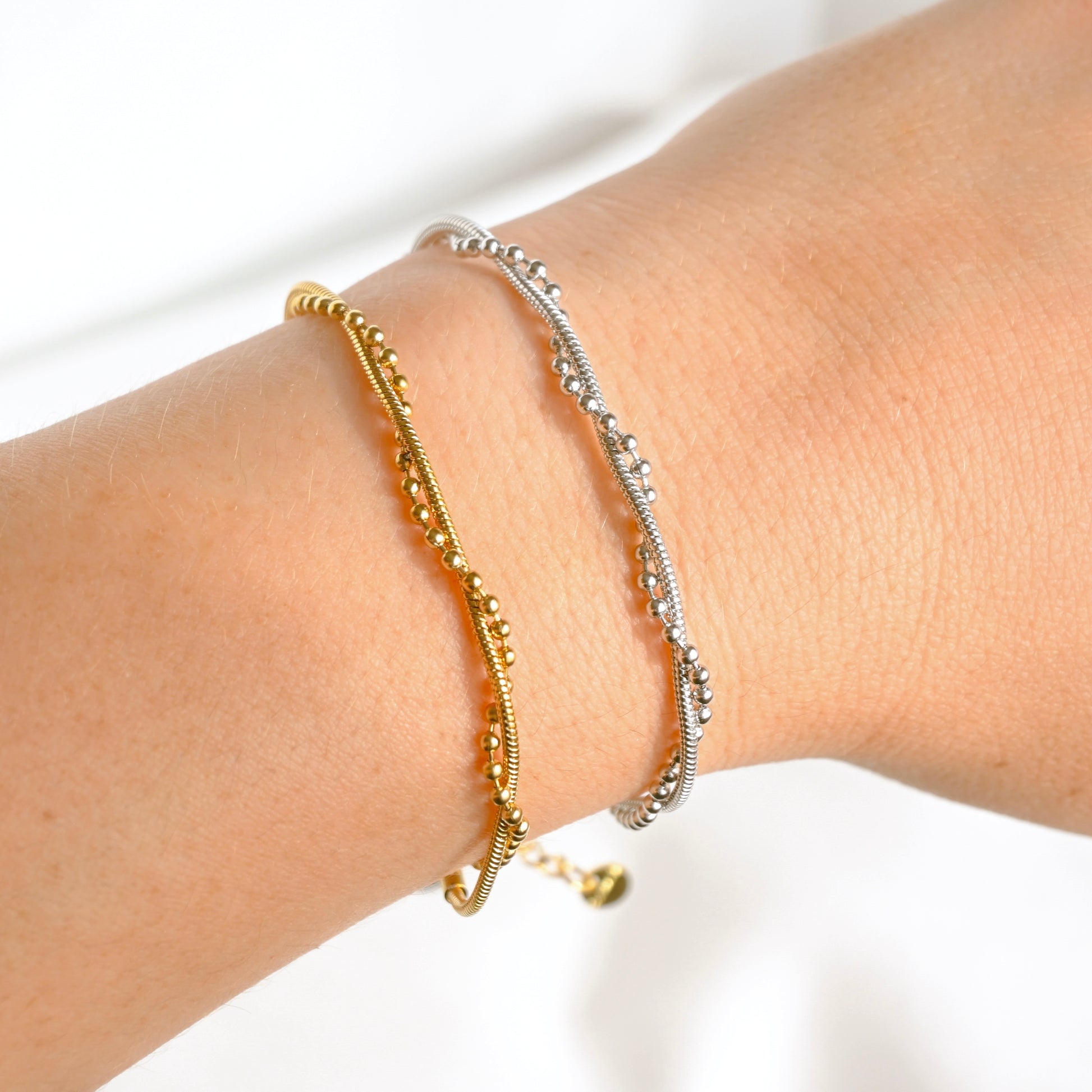 Two delicate twisted beaded and snake chain bracelets, one in yellow gold and one in silver, shown on a person's wrist against a bright white background.