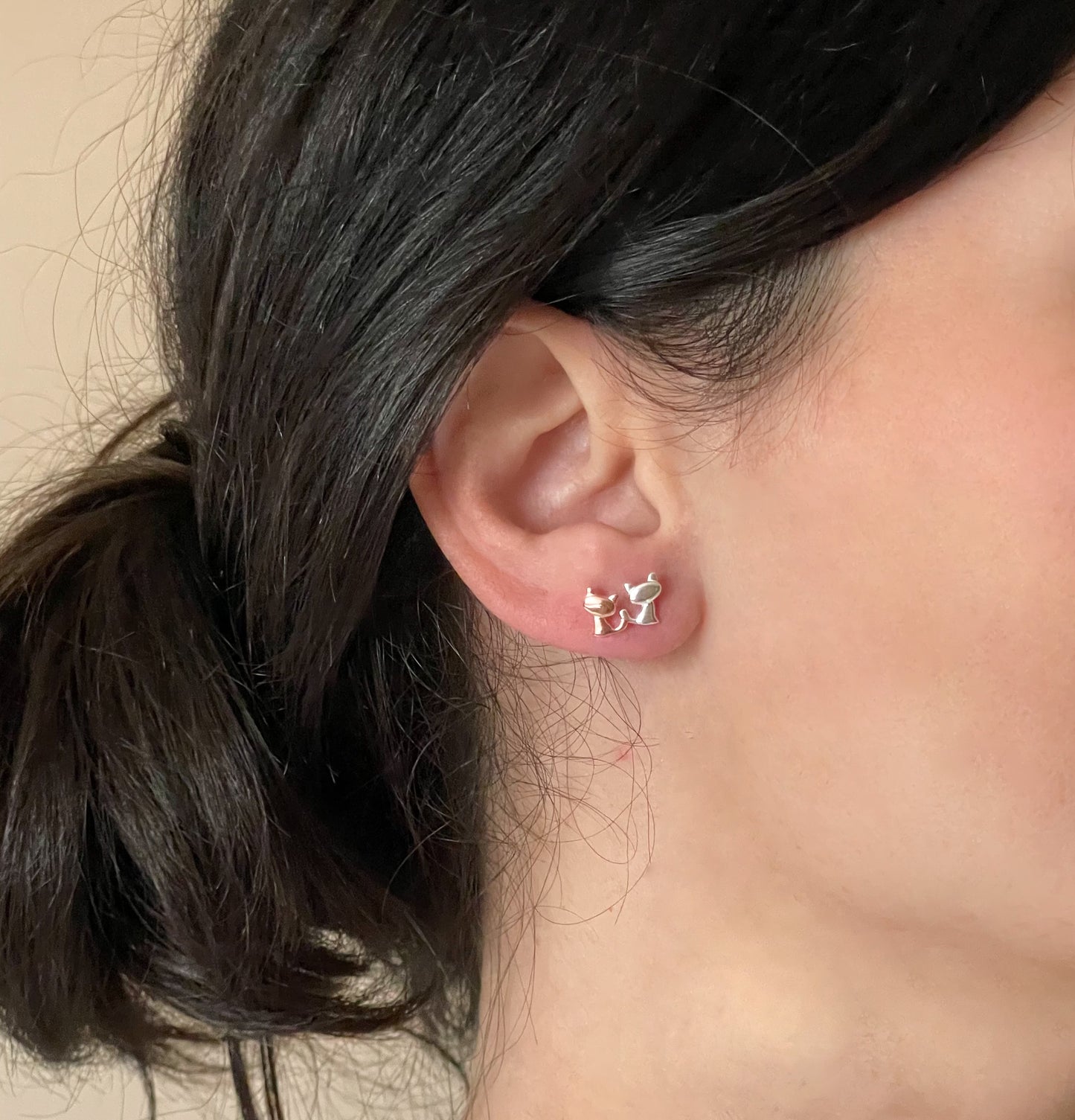 Close-up of an ear wearing two small sterling silver cat earrings, one in silver one in rose gold, with a neutral background