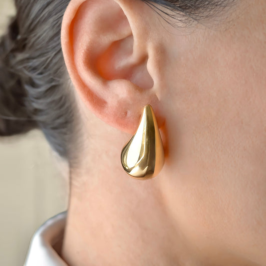 Oversized Gold teardrop earring worn on a person's ear with a neutral background