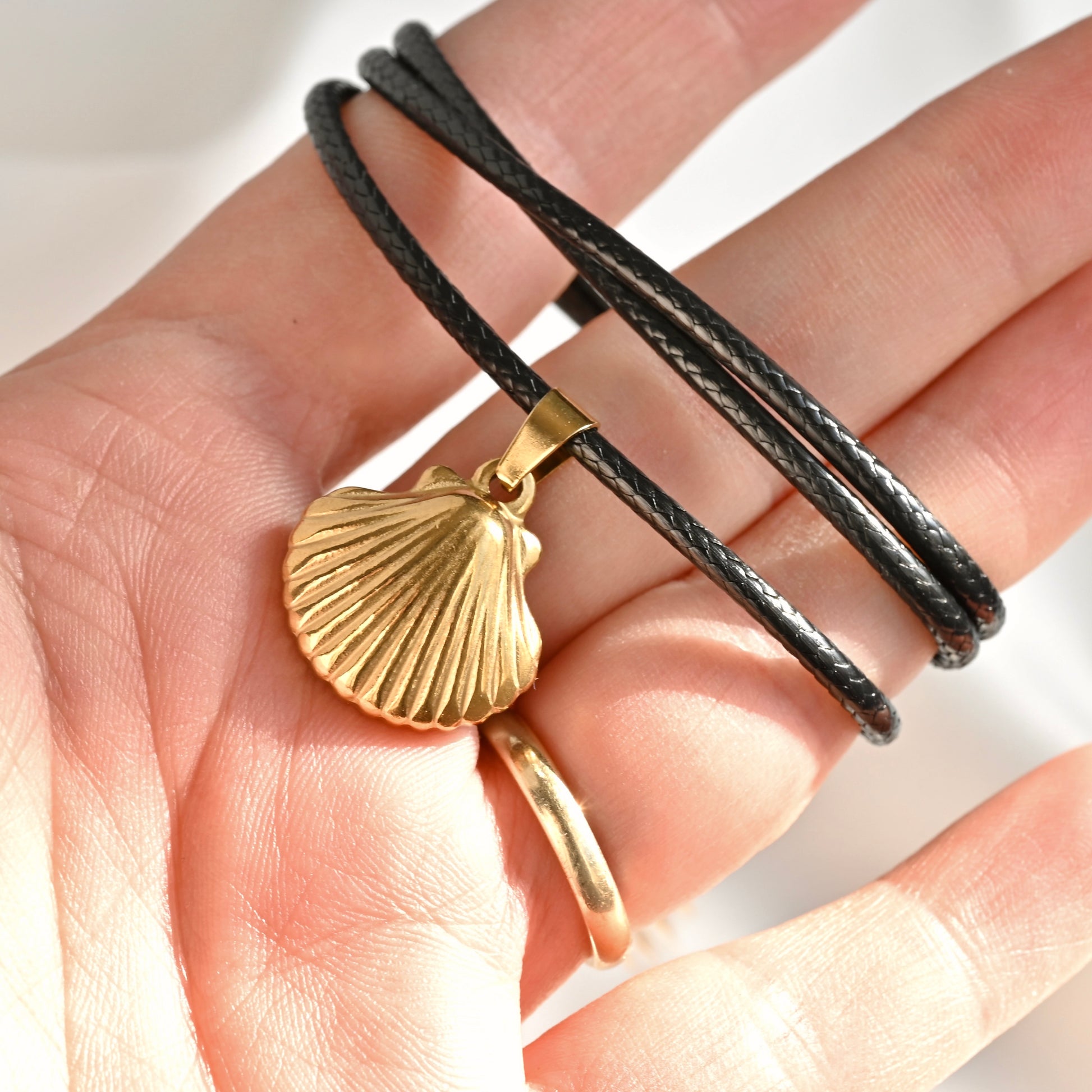 A close-up of a hand holding a black cord necklace with a gold scallop shell pendant, alongside a matching gold ring.