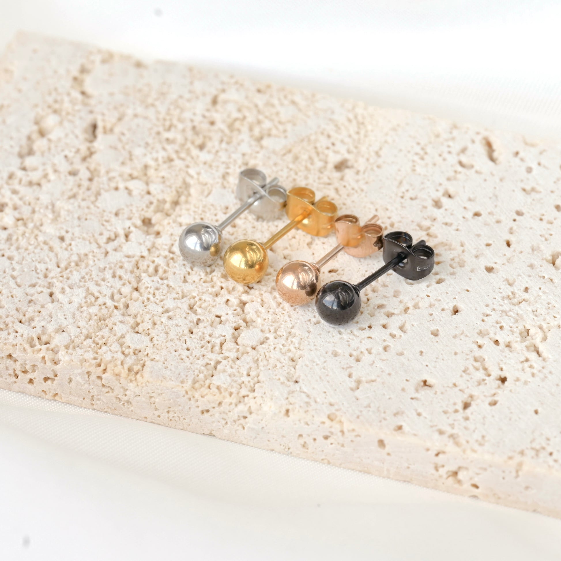 Set of four small spherical stud earrings in silver, gold, rose gold, and black on a textured beige surface.