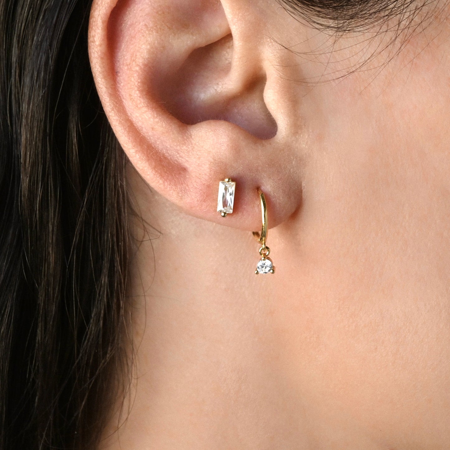 Close-up of a person wearing gold hoop  dangle earrings with clear round CZ stone, stacked with a diamond bar stud in the second hole.