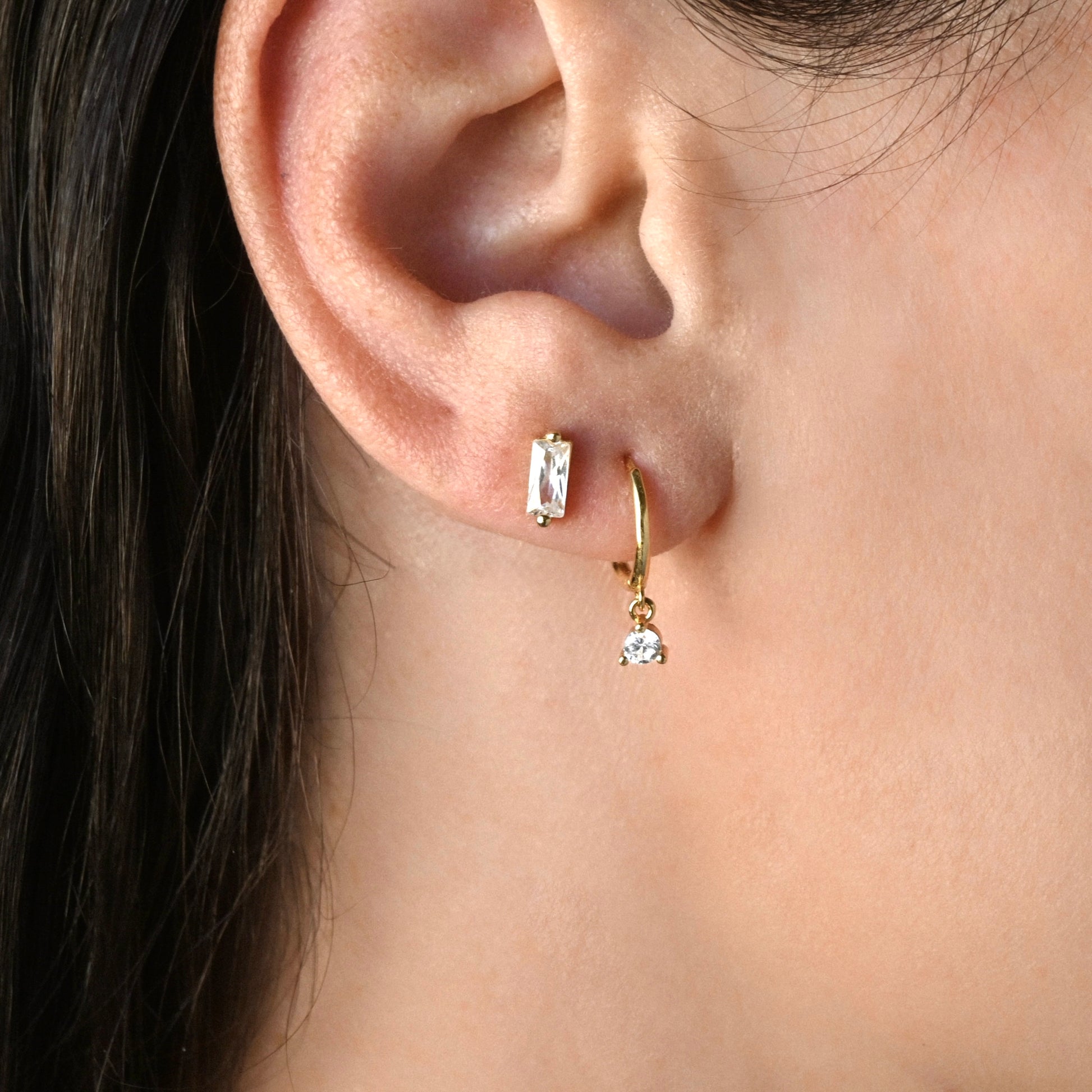 Close-up of a person wearing gold hoop  dangle earrings with clear round CZ stone, stacked with a diamond bar stud in the second hole.