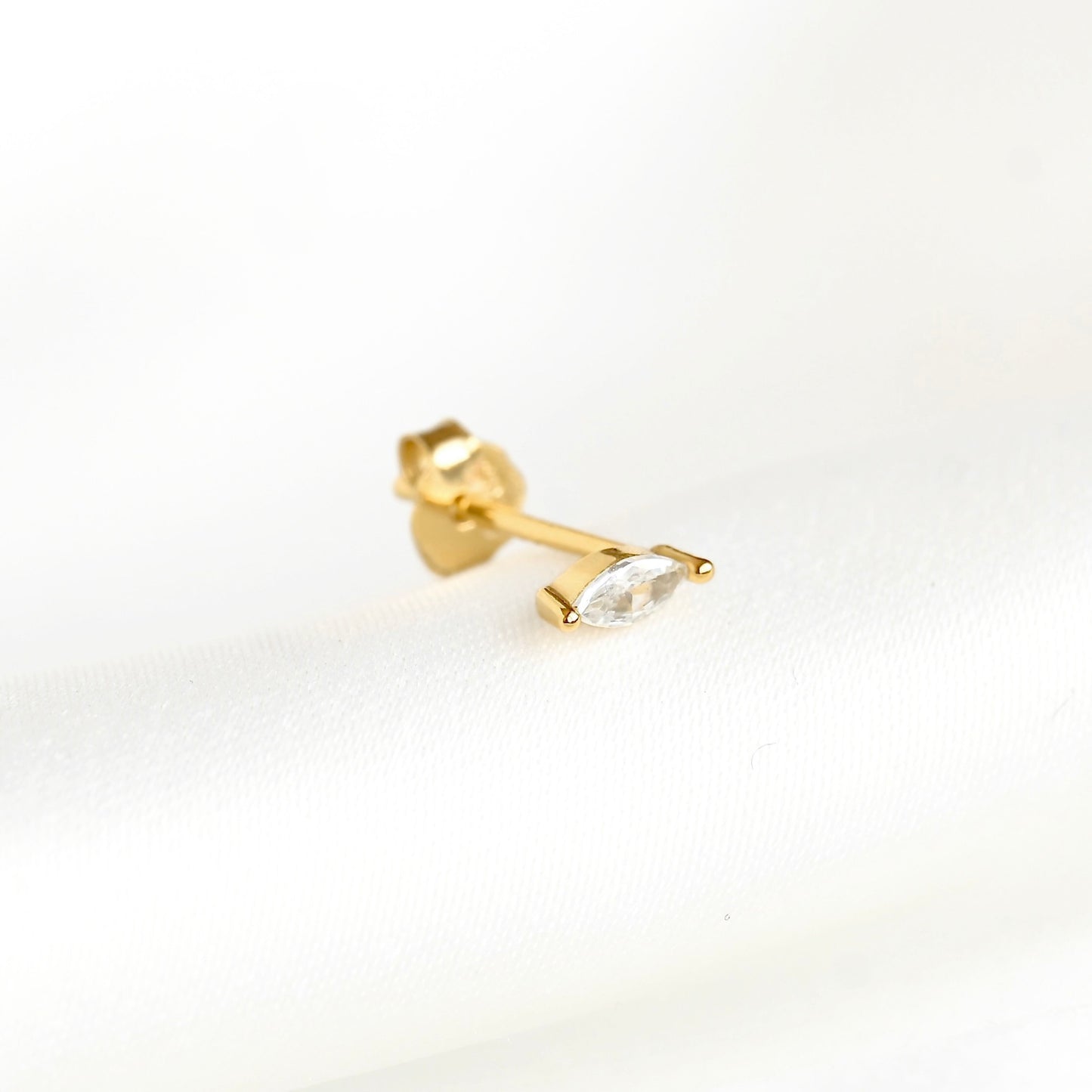A single gold stud earring featuring a marquise-cut clear gemstone, shown on a clean white textured background.