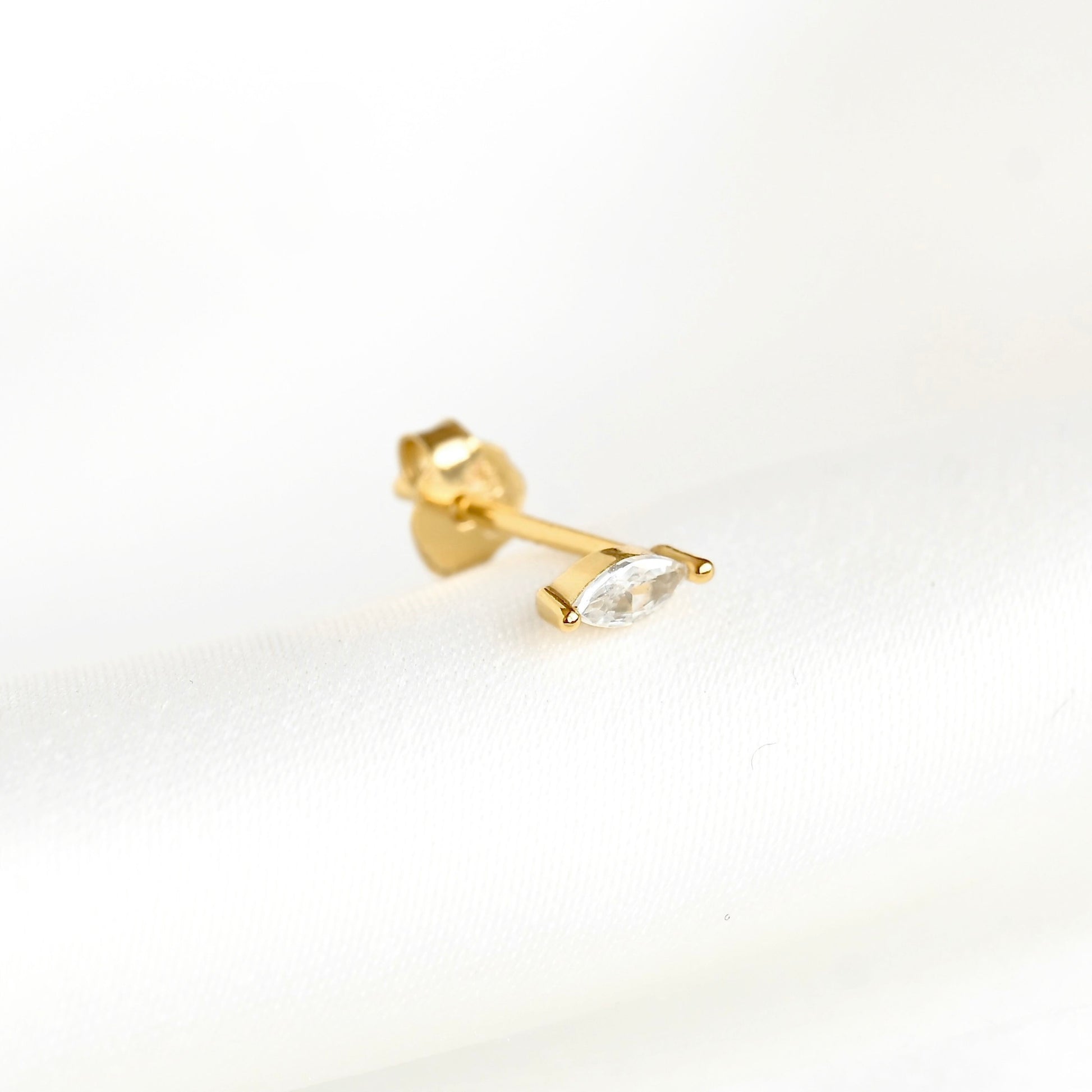 A single gold stud earring featuring a marquise-cut clear gemstone, shown on a clean white textured background.