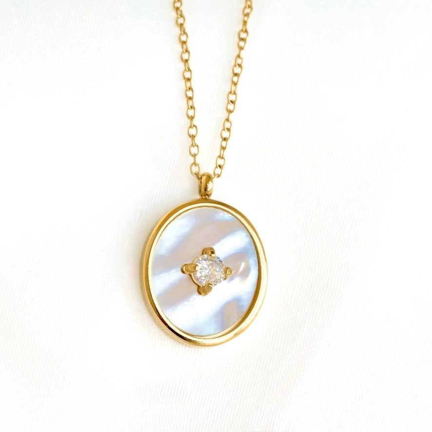 Gold oval pendant necklace featuring a white mother-of-pearl inlay and a small central clear gemstone, shown on a white textured background.