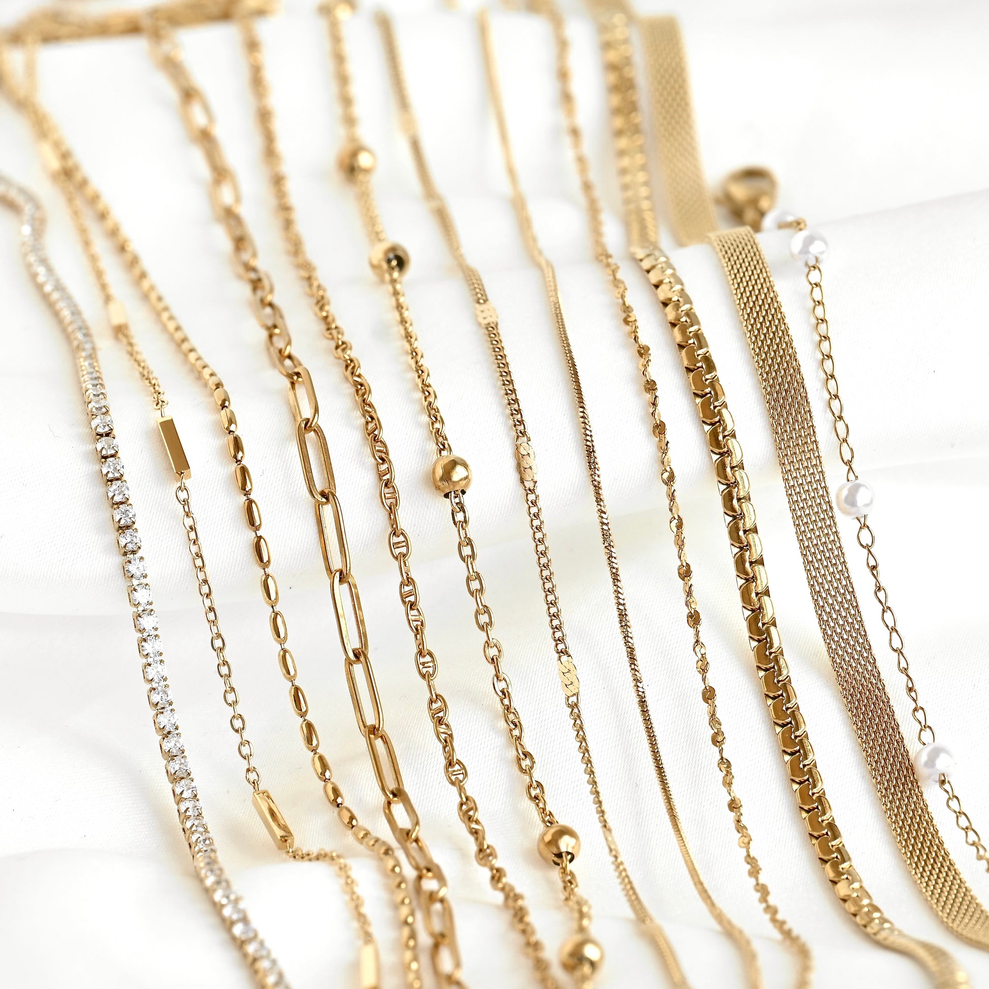 A close-up arrangement of multiple gold-tone chain necklaces with different link styles, displayed on a white fabric background.