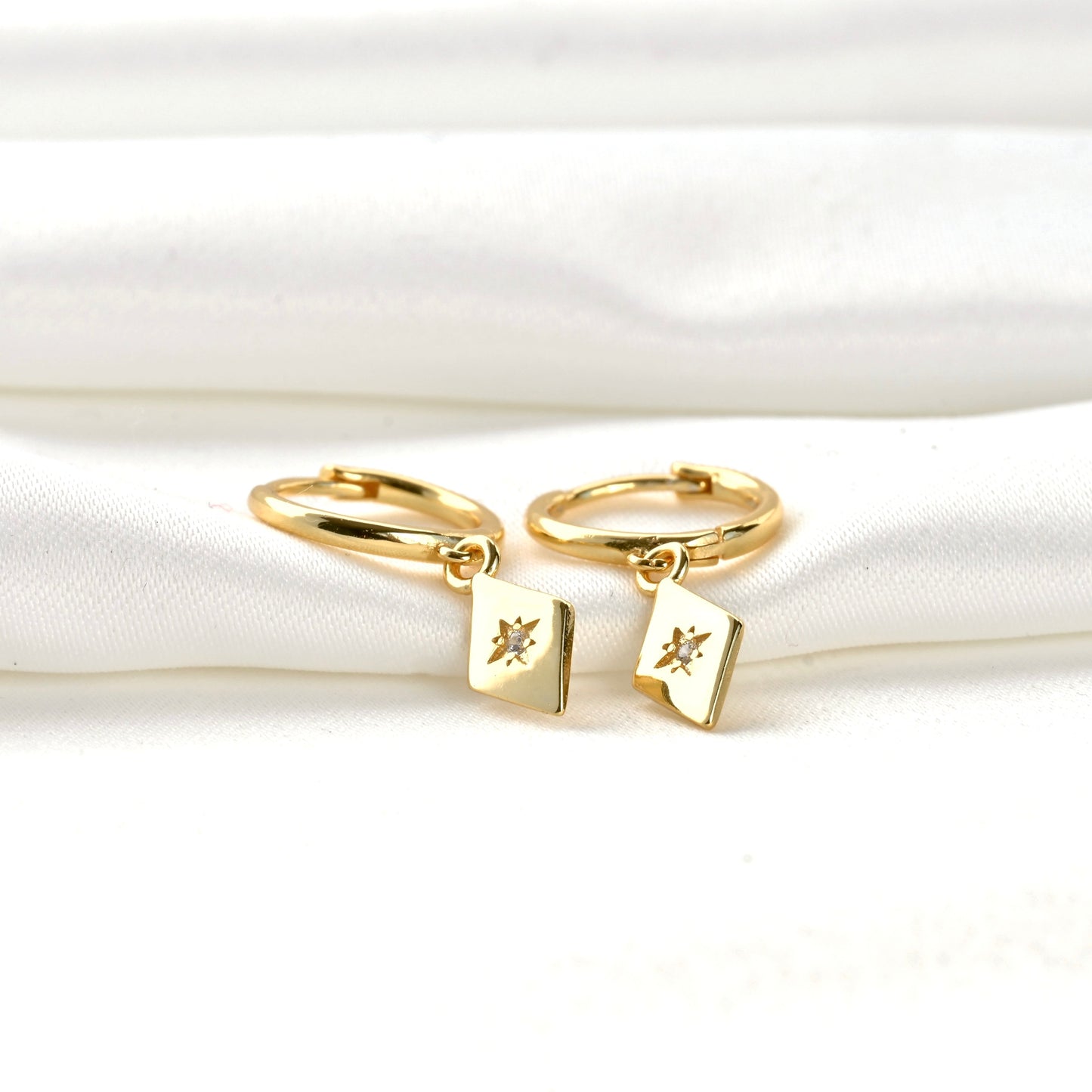 Gold hoop earrings with rhombus-shaped charms on a white fabric background