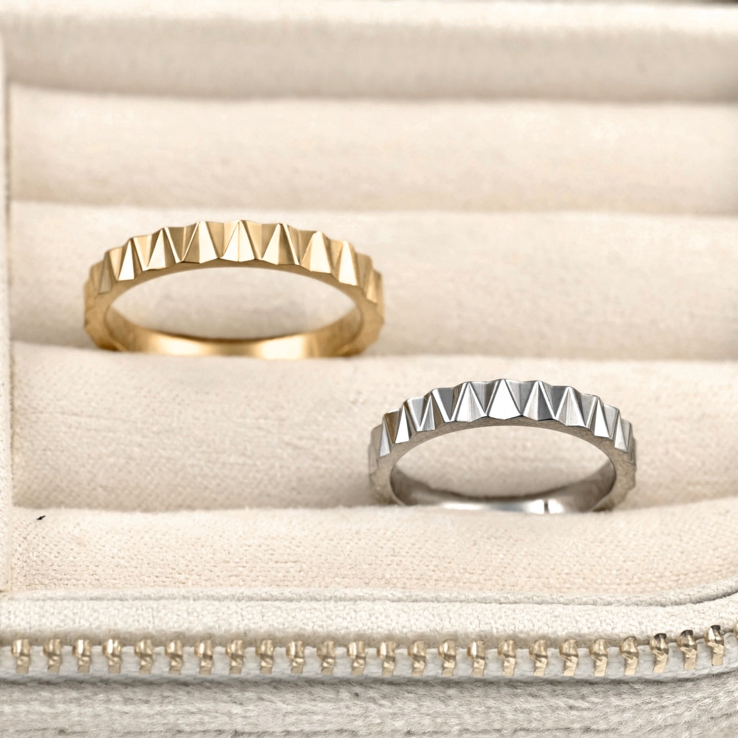 Two geometric pyramid-studded rings, one gold and one silver, displayed on a beige velvet ring box.