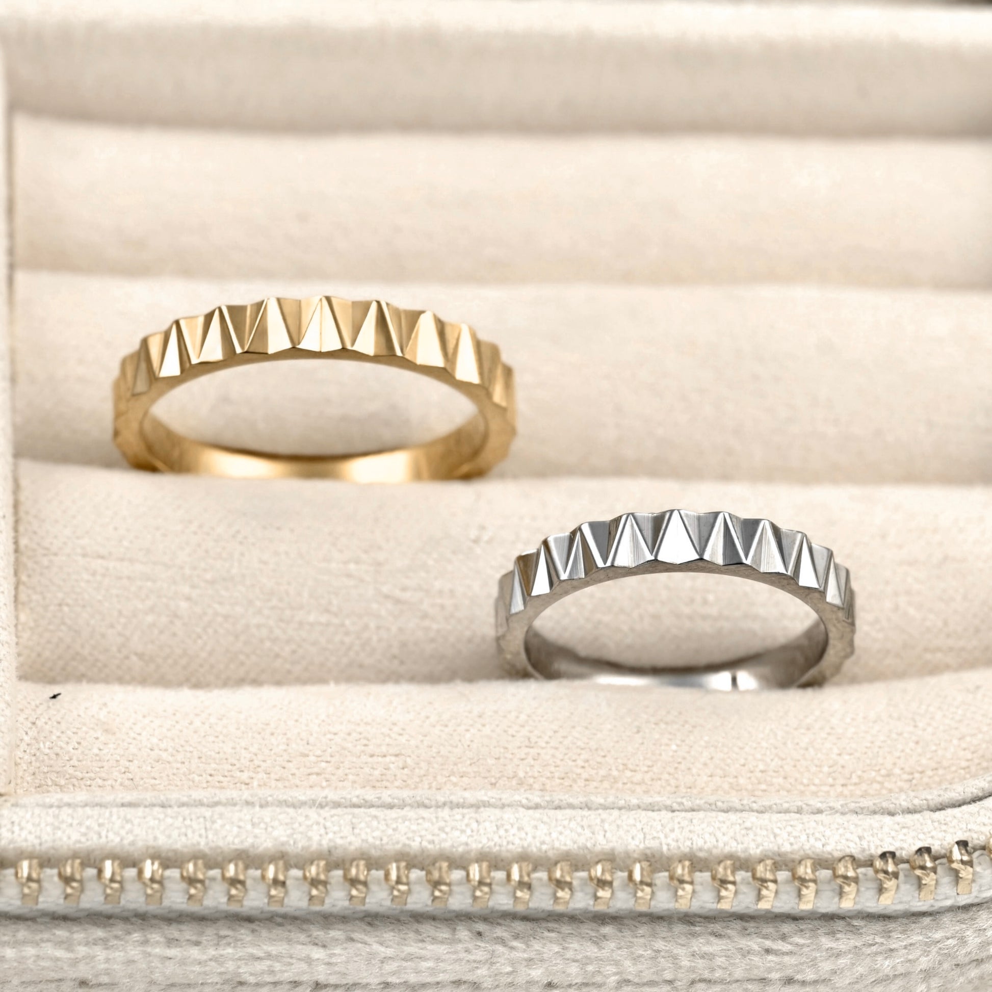 Two geometric pyramid-studded rings, one gold and one silver, displayed on a beige velvet ring box.