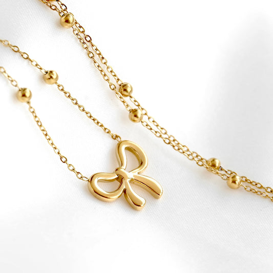 A gold necklace with a bow-shaped pendant and small bead accents on a delicate chain, displayed on a clean white fabric background.