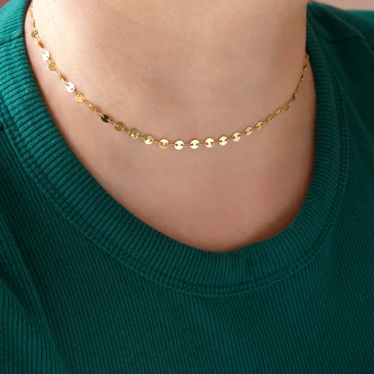 A close-up of a person's neck wearing a delicate gold chain necklace with small flat circular discs, paired with a dark green ribbed knit top.