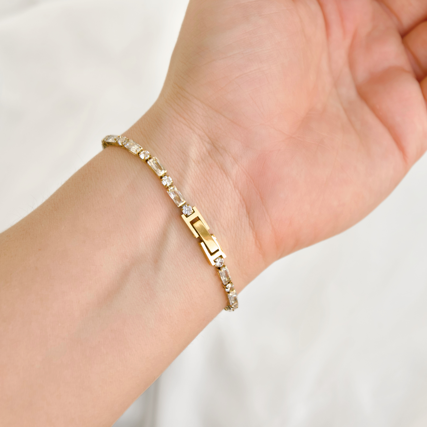 A gold tennis bracelet with clear rectangular and round CZ stones worn on a person's wrist, shown against a soft white background.