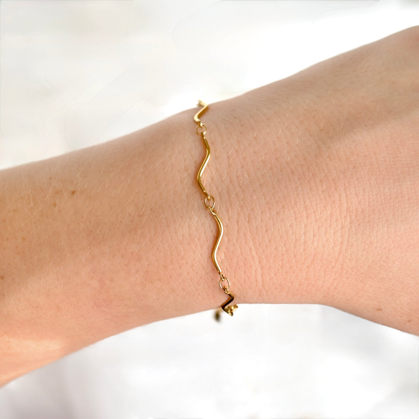 A delicate gold wavy chain bracelet worn on a person's wrist, shown against a soft, bright white background.