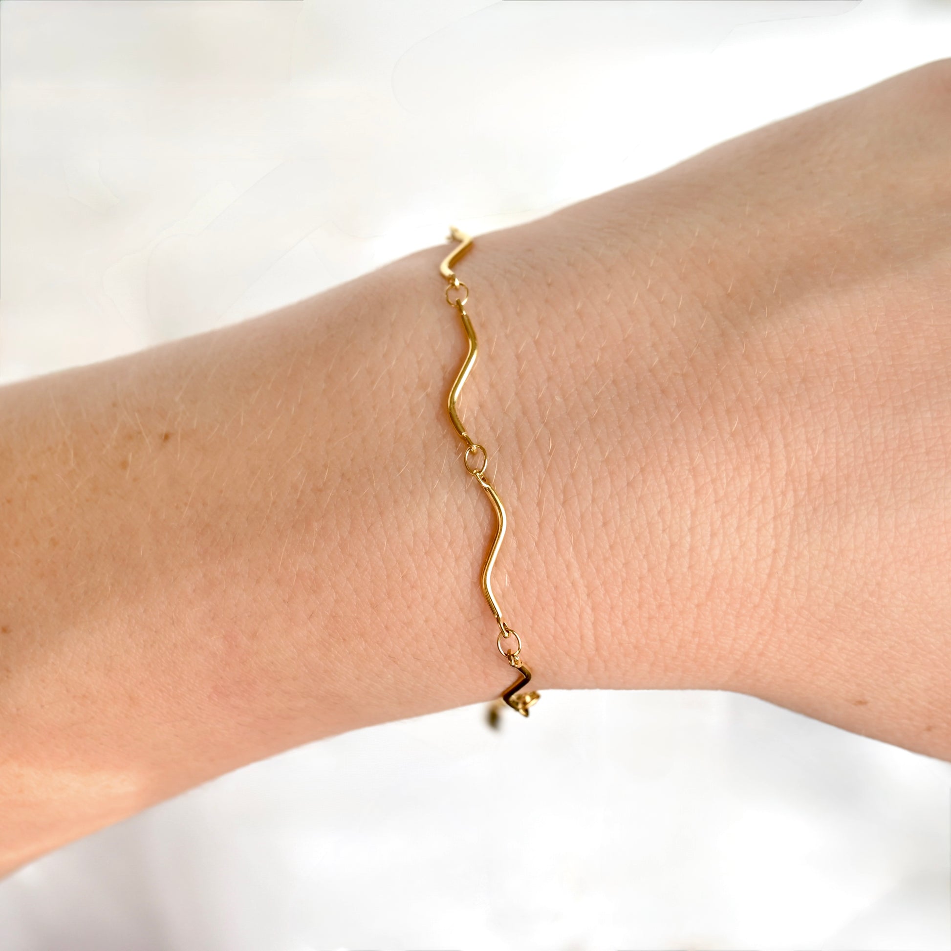A delicate gold wavy chain bracelet worn on a person's wrist, shown against a soft, bright white background.