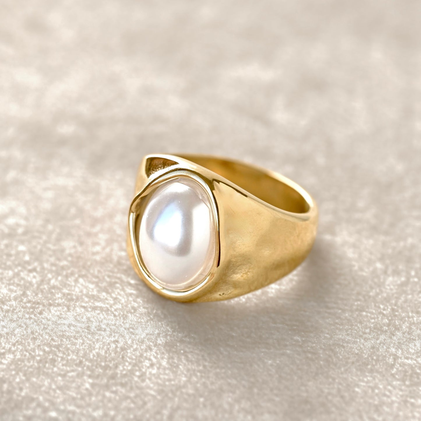 Close-up view of a gold pearl ring featuring a large, smooth white pearl-like cabochon, photographed on a soft beige  velvet  fabric background.