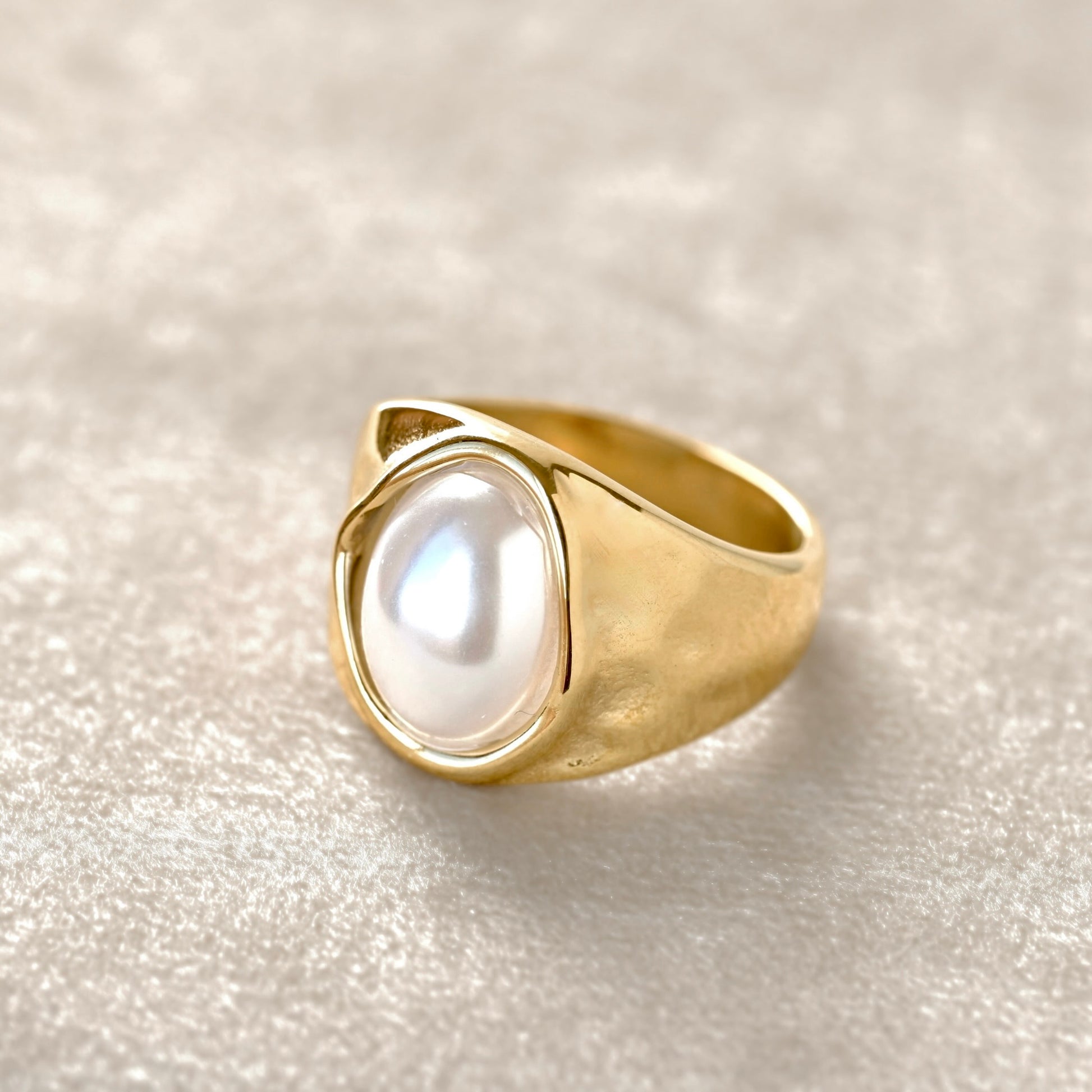 Close-up view of a gold pearl ring featuring a large, smooth white pearl-like cabochon, photographed on a soft beige  velvet  fabric background.