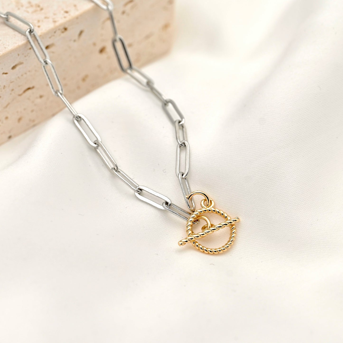 A silver paperclip chain bracelet with a gold-toned toggle clasp pendant, styled on a soft white fabric background with a textured stone prop.