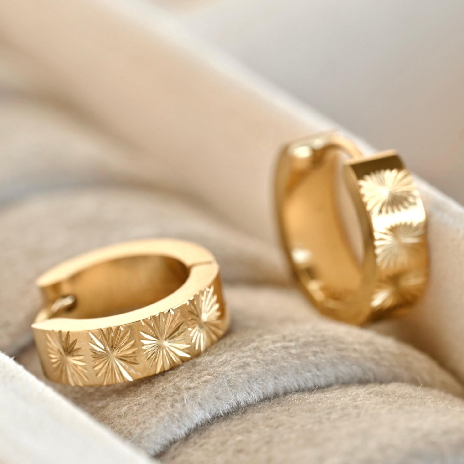 A pair of gold huggie hoop earrings with a sunburst star pattern, resting on a soft beige velvet-lined jewelry box.