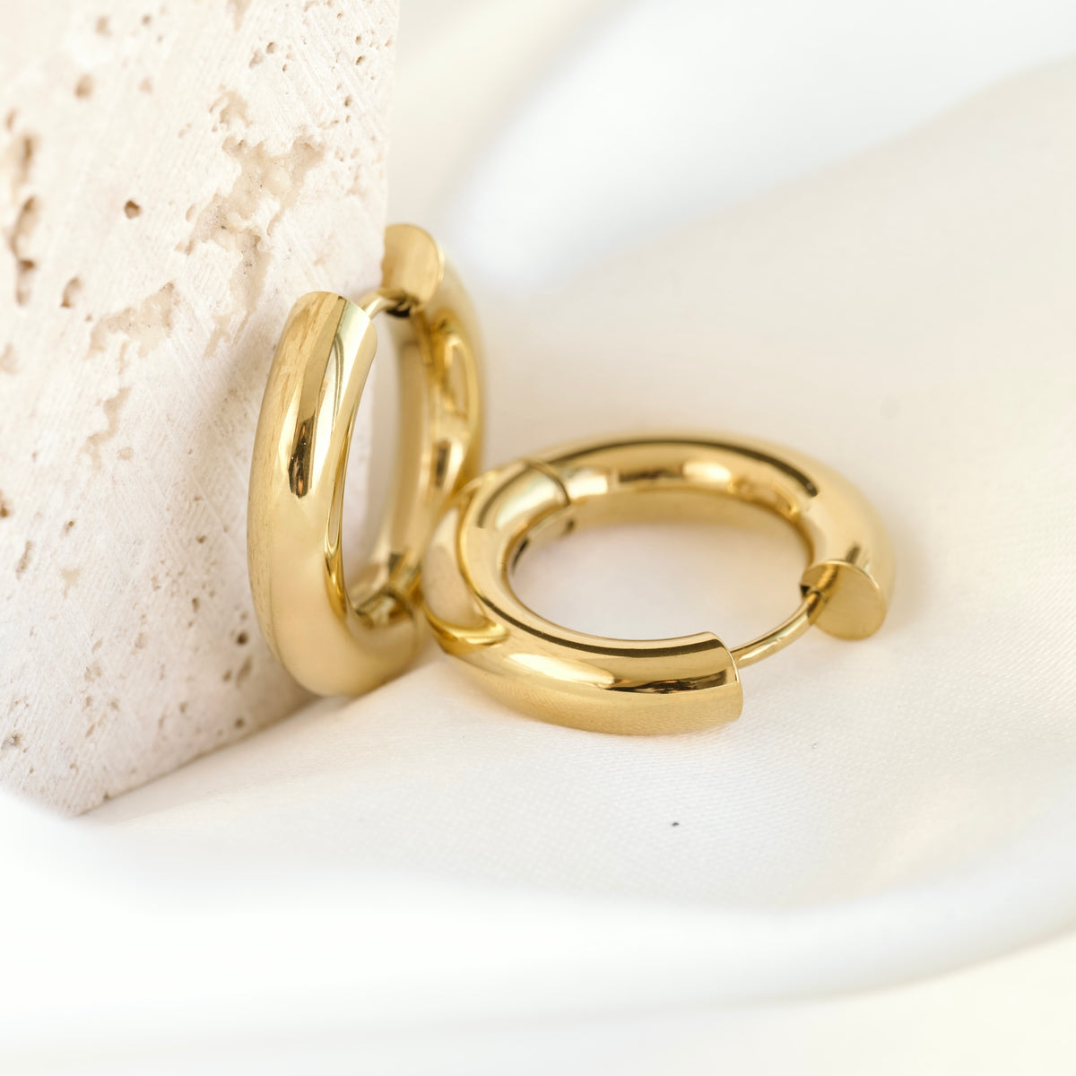 A pair of bold huggie earrings one on white satin the other resting against beige stone.