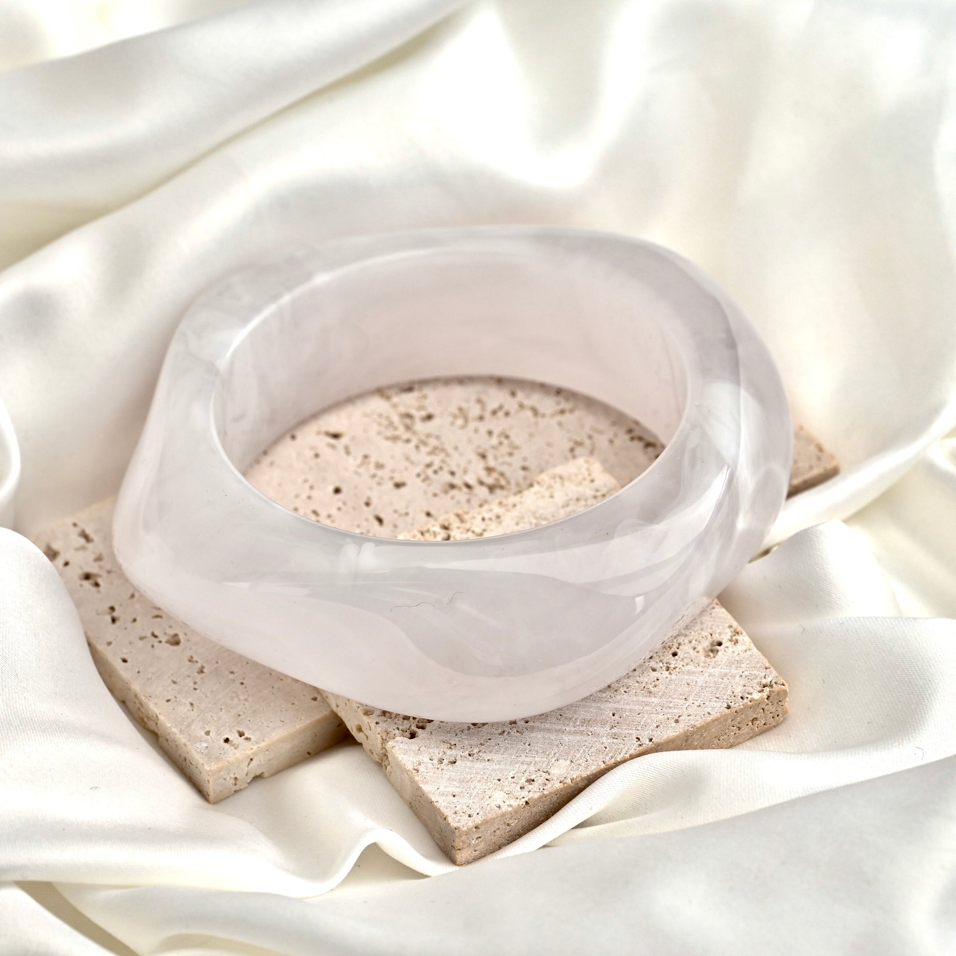 A thick translucent cool white bangle bracelet displayed on a textured stone block over draped white satin fabric.