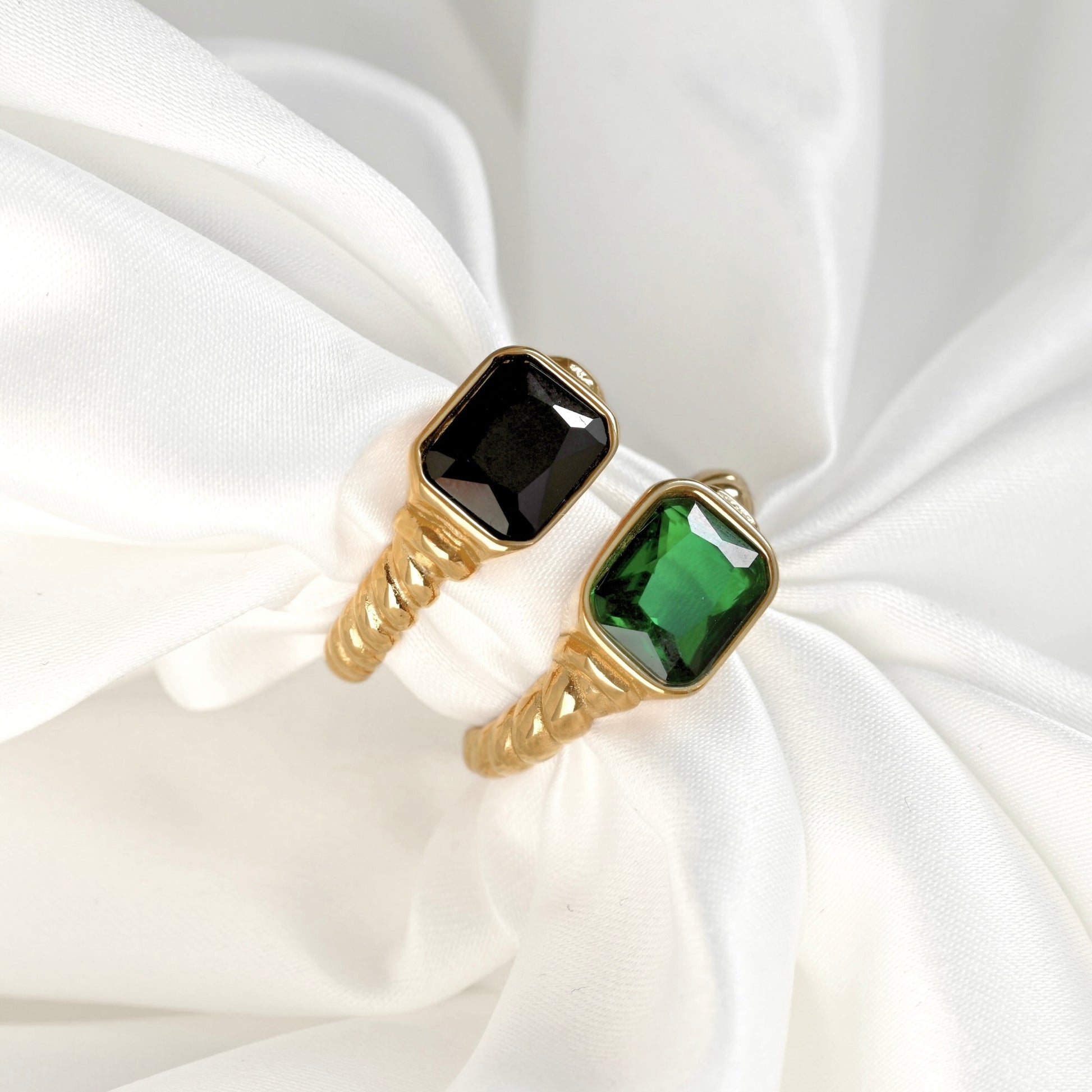 Two gold-plated rings with rectangular faceted gemstones, one black and one green, displayed on a draped white satin fabric.