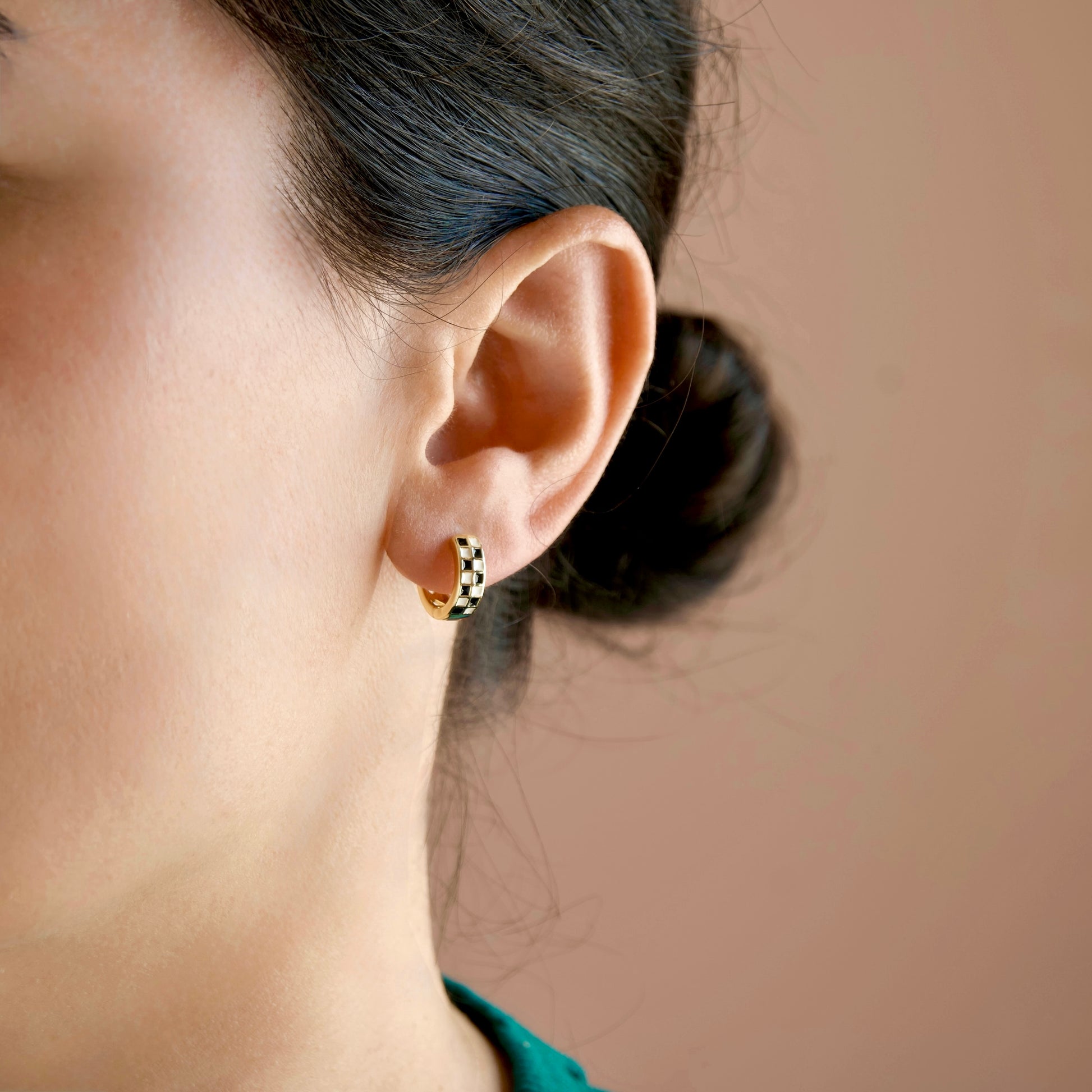 A close-up side profile of a person wearing a small gold huggie hoop earring with a black and white checkered pattern, set against a solid warm beige background.
