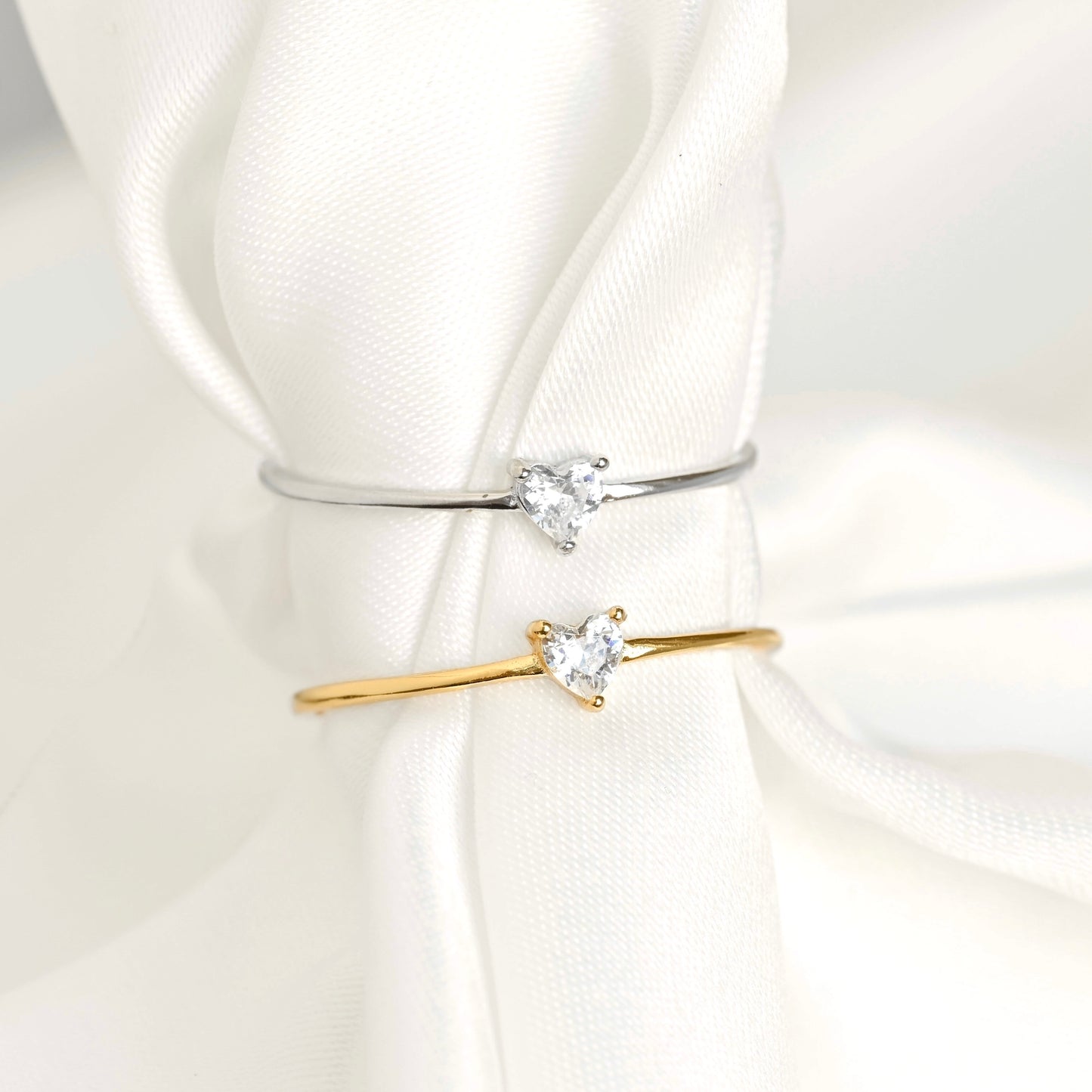Two slim rings with heart-shaped clear gemstones, one in silver and one in yellow gold, displayed on a white fabric napkin.