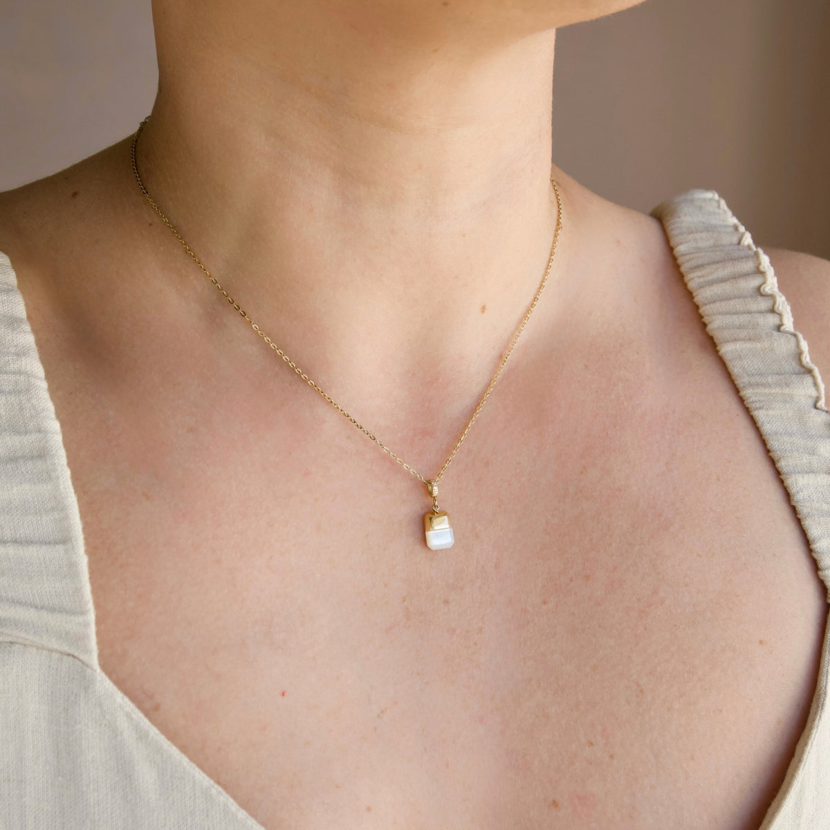 A close-up of a model's neck and chest wearing a delicate gold chain necklace with a small  square pendant featuring a gold top and pearl bottom.